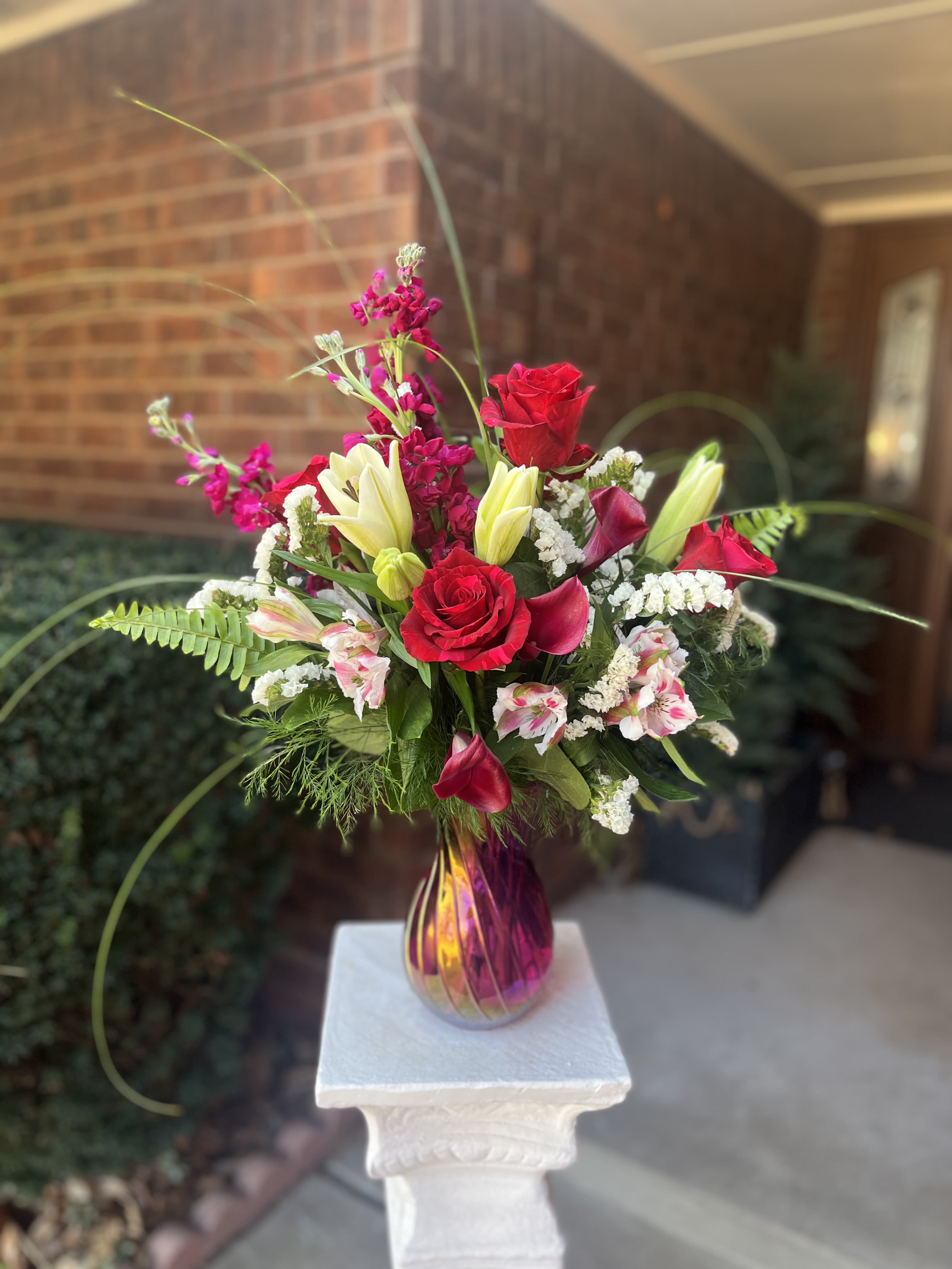 Elegance - A lush cluster of red roses anchors this romantic bouquet, the kind of birthday bouquet or anniversary roses that makes the person you're thinking of feel adored. Vertical and slightly airy, the design pairs the roses with pale yellow-white lily buds, deep pink stock, soft pink alstroemeria and small white filler blooms to create a rich, celebratory mix perfect for romantic celebrations, birthdays, or heartfelt appreciation.  Set in a tall iridescent magenta-and-gold glass vase on a white pedestal, the arrangement reads like a jewel-toned statement piece-glowing from every angle and adding height to tabletops or entryway consoles. Textures range from the velvety rose petals to the spiky, fragrant stock and the sculptural mini calla lilies, giving visual depth and a touch of sweetness in scent. The color palette of red, pink, white and yellow keeps the look warm and affectionate without feeling fussy.  This is the kind of bouquet that makes someone feel truly seen-bold, tender, and celebratory all at once. Order now to send an immediate message of love or thanks; the tall vase presentation arrives ready to display, making it an effortless gift for the person who deserves something memorable.