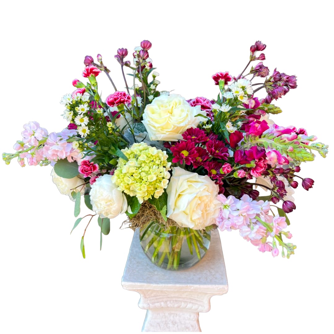 Trinity - A garden-fresh celebration bouquet arranged to make someone feel seen: Trinity is a wide, spreading flower arrangement perfect for birthdays, congratulations, or simple well-wishes. The mix of white and cream roses, cream peonies, with pale green hydrangea gives an elegant, uplifting core, while bright pink and burgundy clusters-magenta chrysanthemums and pink carnations-bring warmth and depth that read like a joyful embrace.  This design pairs softer pastel pink stock and small white daisy-like blooms with taller snapdragon stems and textured chrysanthemums for a natural, layered silhouette. Displayed in a clear round glass vase, the gathered stems are visible for an organic, relaxed look; the arrangement sits on a white pedestal stand that adds a clean, celebratory lift. Colors include pink, white, green, and touches of red/burgundy for contrast.  Ideal for sending appreciation, celebrating milestones, or brightening someone's day, Trinity feels like a thoughtfully gathered garden in bloom-the kind of bouquet that makes the recipient feel genuinely celebrated. Vase included; keep flowers in fresh water, trim stems and remove foliage below the waterline when changing water to extend vase life.