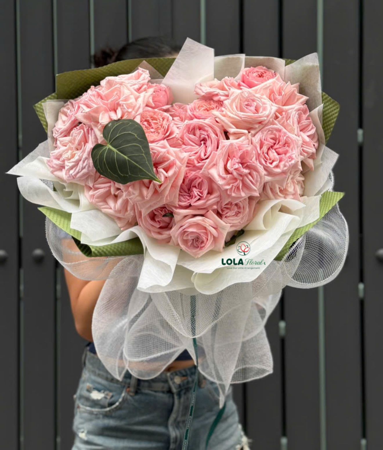 Pink Rose Heart - A dense heart of soft pink roses creates an unmistakable romantic focal point in this flower arrangement-perfect as an anniversary bouquet, Valentine's gift, or a heartfelt wedding gesture. The heart-shaped cluster reads immediately as an expression of love and tenderness, the kind of gift that will make the person you're thinking of feel truly cherished.  Tightly packed pink roses form the sculpted heart, their velvety petals contrasted by a single dark, heart-shaped leaf for visual depth. The blooms are framed with crisp white paper and airy white mesh, backed by green paper for a fresh pop of color, and finished with a neat ribbon tie. The overall palette of pink, white, and green feels modern and elegant while still intimate.  Choose this arrangement when you want to send a clear message of affection-whether marking a milestone, surprising a partner, or offering a romantic birthday bouquet. It arrives ready to display and looks best in a low, wide vase; keep roses in fresh water, trim stems slightly, and refresh the water daily to extend the life of the blooms.