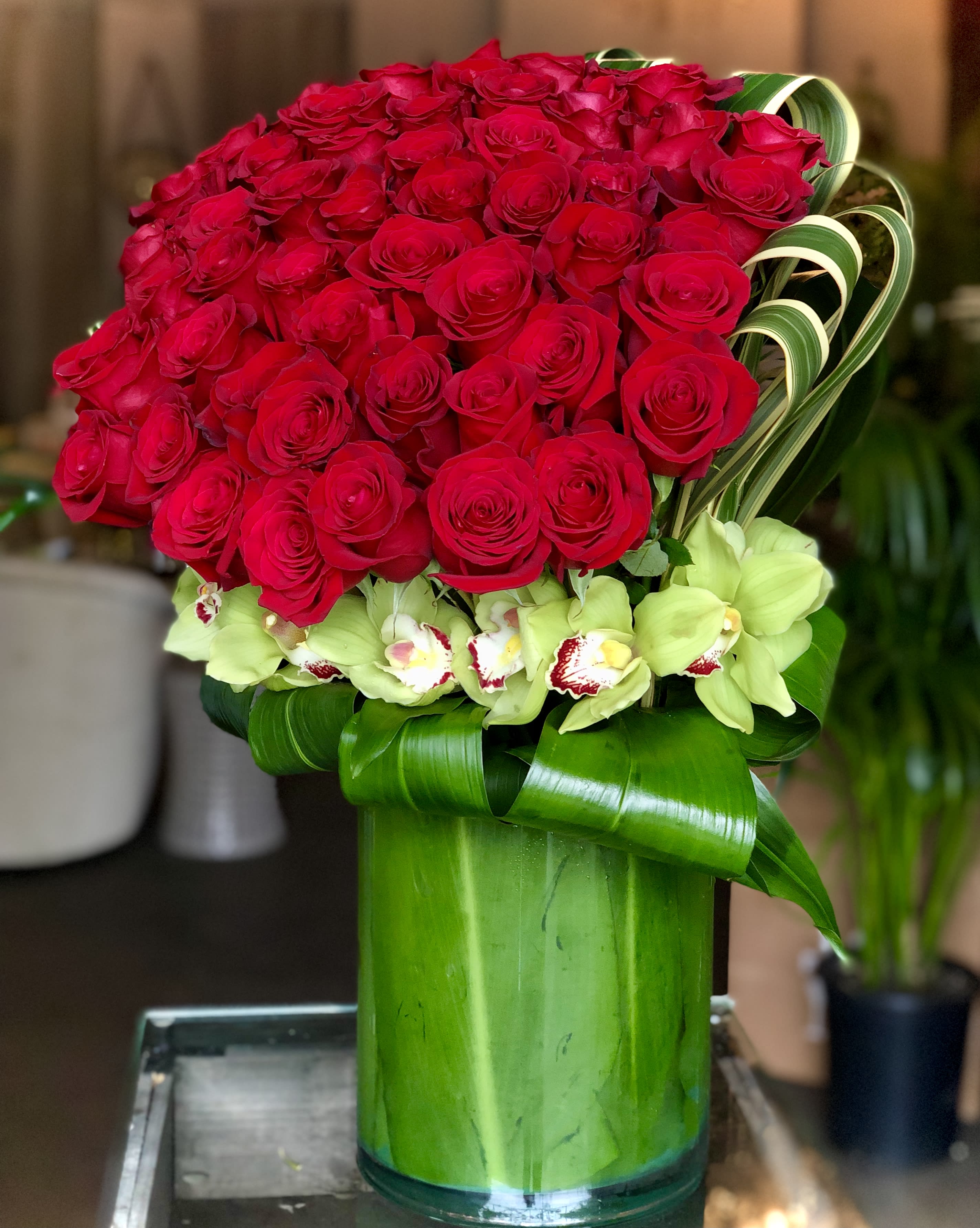 Tall arrangement of red roses with green cymbidium orchids in a leaf-lined green glass vase