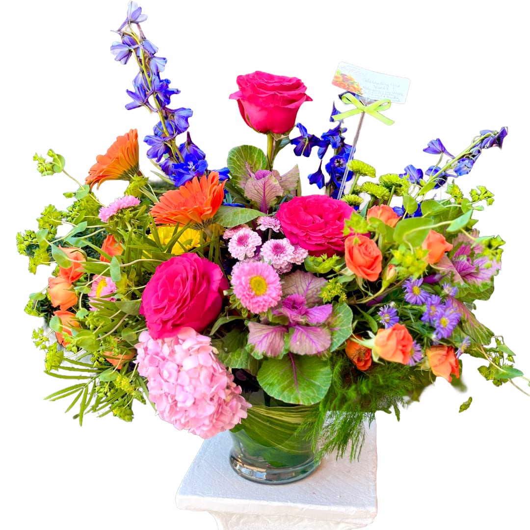 Celebration - A bold burst of hot pink roses and electric orange gerbera daisies greets the eye-this Celebration flower arrangement is a birthday bouquet and congratulations bouquet that reads joyful from across the room. Tall blue‑purple delphinium spikes and vivid purple asters lift the composition, while lush pink hydrangea and bright green foliage soften the shape so the person you're sending it to feels truly celebrated and seen.  Densely packed for a full, rounded effect, the design mixes roses, gerbera, hydrangea, delphinium, asters and button chrysanthemums with textural accents like ornamental cabbage and variegated leaves. Available in a clear glass vase or a clear glass cylinder vase with broad leaves wrapped inside to hide stems, each arrangement includes a card pick with a small card and ribbon for your personal message. The saturated pinks, oranges, purples and greens make this an uplifting choice for birthdays, congratulations, Mother's Day or get-well wishes.