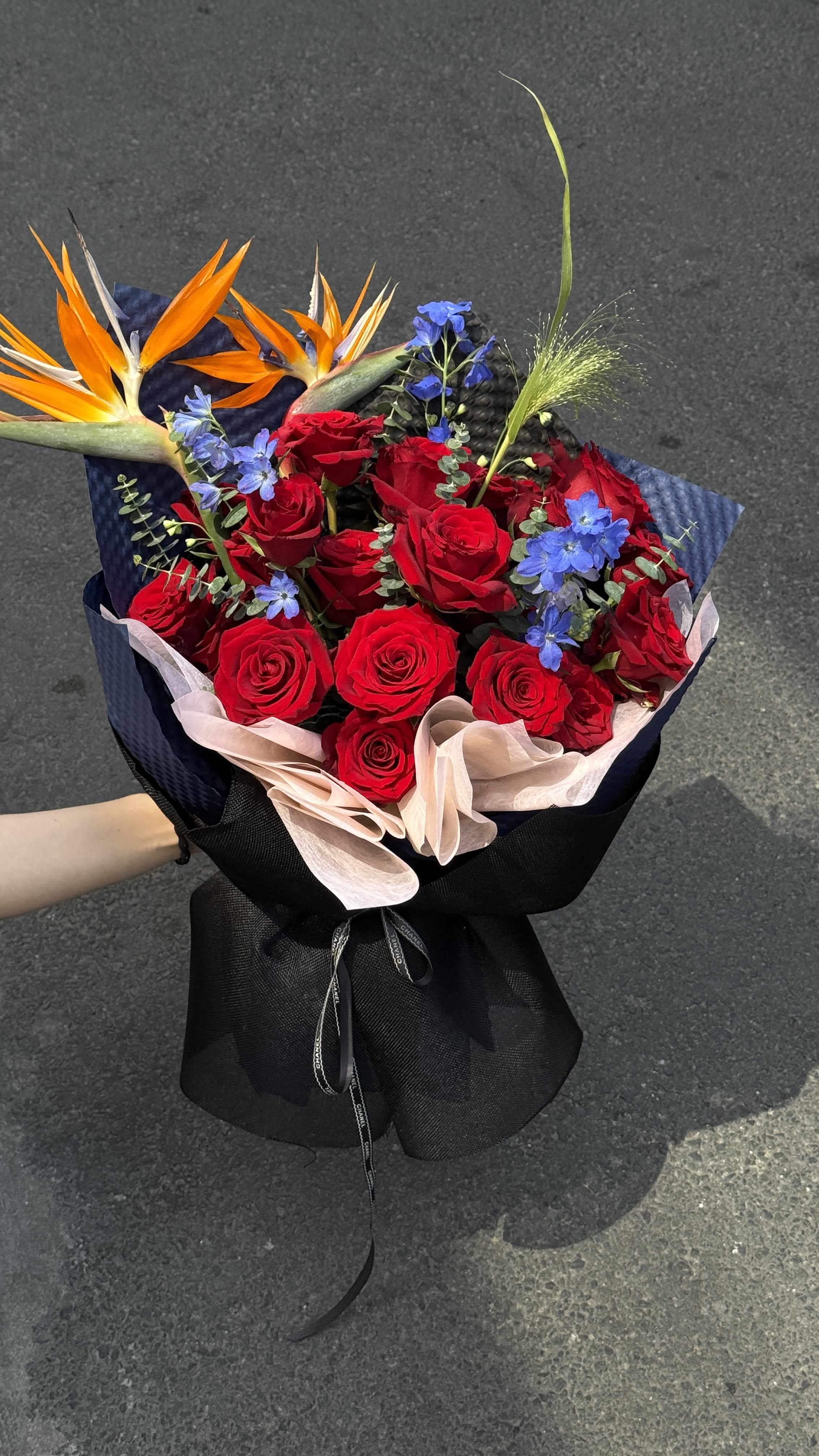 Hand Wrapped Bouquet 02 - A dense cluster of vivid red roses anchors this hand wrapped bouquet, instantly signaling romance and celebration - perfect as an anniversary roses bouquet or a dramatic birthday gift for someone you adore. The vivid red blooms are offset by small blue filler flowers and two bright orange bird-of-paradise blooms that peek above the roses, creating a bold, tropical contrast that reads both passionate and joyful for special occasions.  Wrapped in layered dark navy and pale blush decorative paper and tied with a large black ribbon bow, the presentation feels luxe and purposeful; the navy grounds the color story while the blush softens the edges. Texturally there's the velvet of the roses, the fine, playful texture of the blue fillers, and the sculptural orange blooms that add height and movement. Sized to make a statement without overwhelming, it's the kind of bouquet that will make the person you're thinking of feel celebrated and seen.  Ideal for anniversaries, romantic surprises, or milestone celebrations, this arrangement arrives ready to display - no extra vase required, though a tall vessel highlights the bird-of-paradise spikes beautifully. To keep blooms looking their best, replace water daily and trim stems at a diagonal before arranging in fresh water.