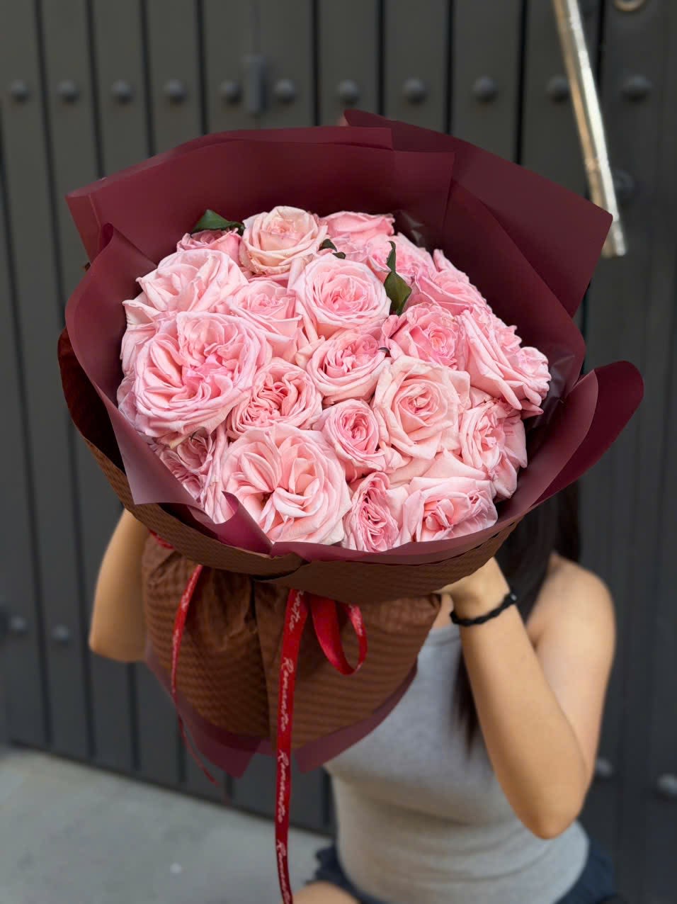 Hand Wrapped Bouquet 03 - A lush cluster of pale pink roses set against deep burgundy wrap makes this romantic bouquet an ideal anniversary roses gift or a love-themed celebration bouquet. The soft blush blooms read tender and affectionate while the rich burgundy paper and red ribbon give the arrangement a dramatic, passionate contrast-perfect for the person you want to make feel adored.  Dense, rounded and generously sized, the hand-held bouquet is made up entirely of pale pink roses for a full, layered texture that looks abundant from every angle. It's finished in decorative burgundy paper, tied with a red ribbon, and can be presented as-is or nestled in the included basket for a more substantial display. The mix of pink, red and white tones reads classic and elegant without feeling fussy.  Give this bouquet to mark anniversaries, romantic milestones, or to say "I'm thinking of you" in a way that feels truly seen; she'll feel cherished the moment she holds it. For best longevity, keep stems in fresh water and trim ends before arranging if transferring from the wrap to a vase.