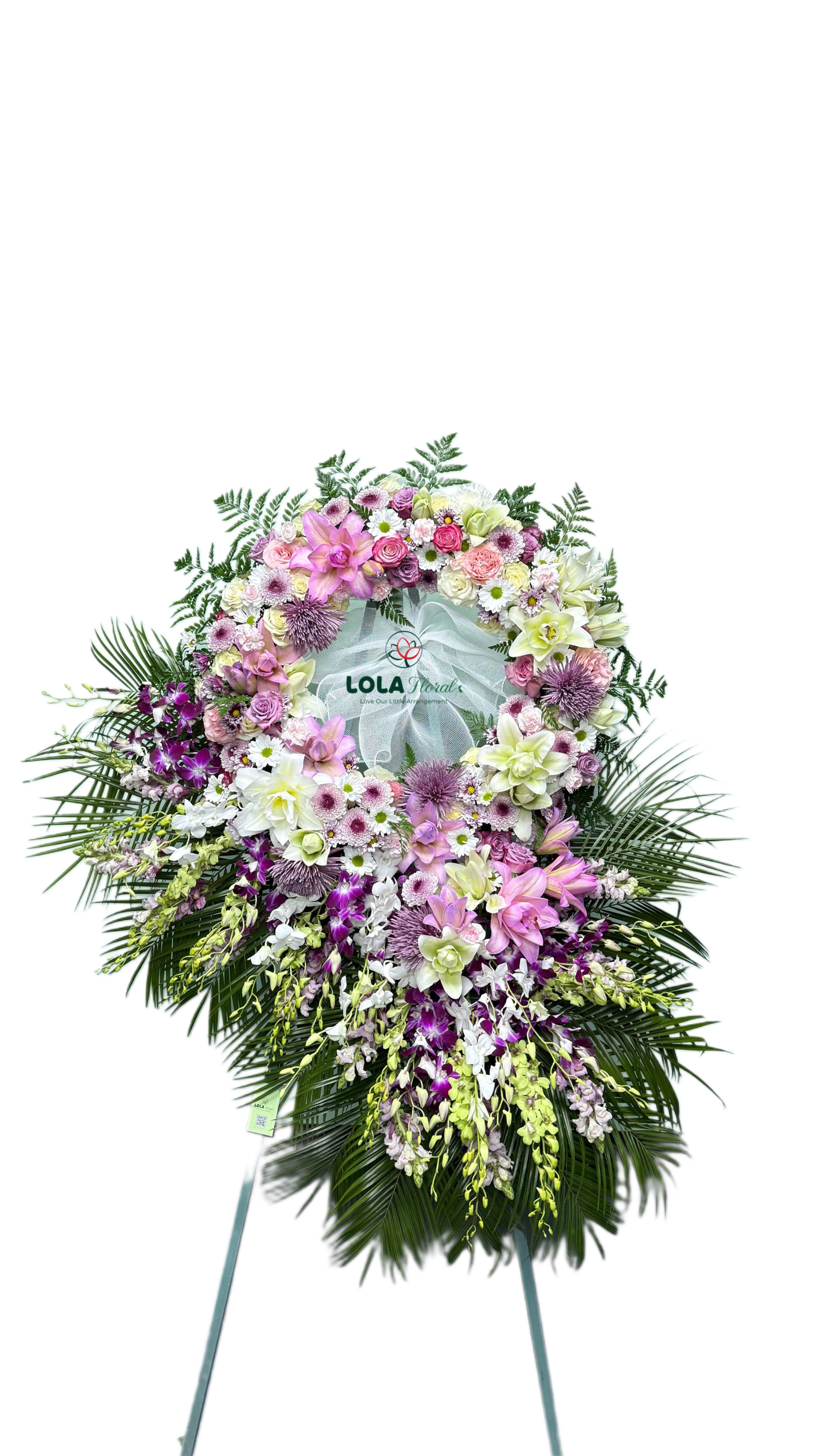 Eternal Peace 03 - A serene circular wreath of pastel pink, lavender, and white blooms makes a striking sympathy standing arrangement for memorial services. The Eternal Peace 03 wreath is designed as a formal tribute that reads gentle and composed from across a room - the person you're honoring will be remembered with a graceful, comforting display.  Densely packed roses, lilies, chrysanthemums, and small daisies form the wreath's textured ring, while sprays of dendrobium orchids cascade down over large palm fronds to add soft vertical movement. A sheer white fabric center and bow soften the composition, with a printed logo card and a sturdy metal easel stand and wreath frame included for easy display. The palette of pink, white, purple, and green keeps the piece elegant and quietly hopeful.  Perfect for funeral services, memorials, or as a sympathy gift to the family, this arrangement conveys respect and calm without being ostentatious. It arrives ready to stand and display; for lasting presentation keep it in a cool, sheltered area until the service and ask our team about local delivery or same-day options when available.