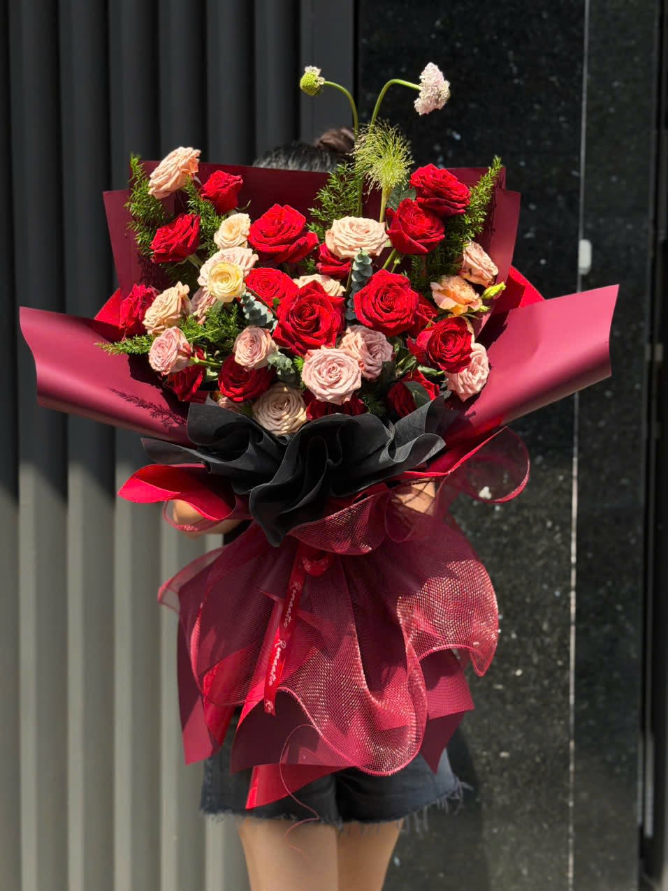 Hand Wrapped Bouquet 04 - A lush cluster of red and blush roses forms the heart of this romantic bouquet, perfect as a Valentine's Day bouquet or an anniversary roses gift that reads as boldly passionate. The rounded, hand-tied style keeps the focus on velvety petals and a warm red-to-pink gradient, so the person you're thinking of will feel genuinely celebrated the moment they see it.  Wrapped in layered burgundy and black decorative paper with a large sheer ribbon bow, this arrangement has dramatic, high-style presence-ideal for someone who loves luxe, contemporary flair. A few airy filler stems and taller accent blooms rise above the main cluster to add movement and a taller silhouette; finished with quality ribbon, it arrives ready to display or hand to a loved one.  Choose this bouquet when you want to make a confident romantic statement-for anniversaries, Valentine's Day, or any moment that calls for something unmistakably special. Hand-tied for easy arranging at home; trim stems and refresh water daily to extend the life of the roses.