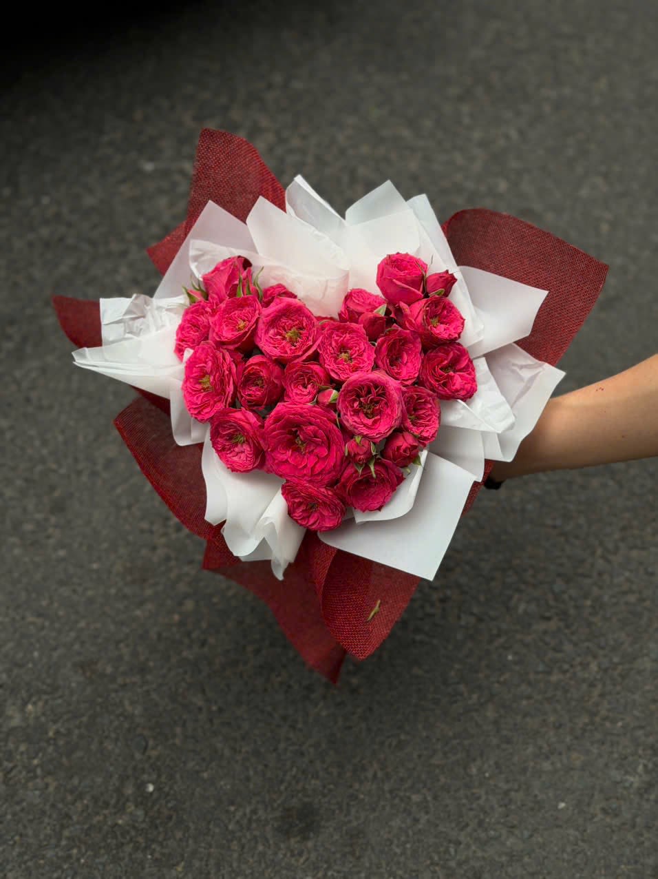 Hand Wrapped Heart Bouquet  - A dense cluster of vivid pink roses arranged into a Hand Wrapped Heart Bouquet that reads as pure romantic intent - perfect as a birthday bouquet, anniversary roses, or a Valentine's Day gift. The compact, hand-tied presentation and heart-like silhouette make the recipient feel truly seen and celebrated, whether you're saying "I love you" or "happy birthday."  Tightly layered pink roses form the visual core, surrounded by crisp white bouquet wrap and a bold burgundy outer paper that frames the blooms with dramatic contrast. The bouquet's compact size and neat hand-wrapped finish give it a polished, gift-ready look; no vase needed if they want to admire it as-is before arranging. Care: keep stems in fresh water, trim ends and recut after a day or two to extend vase life.