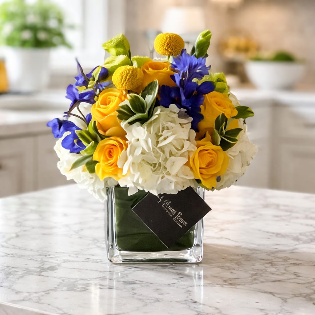 Bluebell - 4X4 glass cube arrangement.  Our bluebell arrangement is a mix of yellow florals with a hint of blue florals  in a clear cube vase with green t leaf lining inside. the perfect arrangment for any occasion to send to your love ones. 