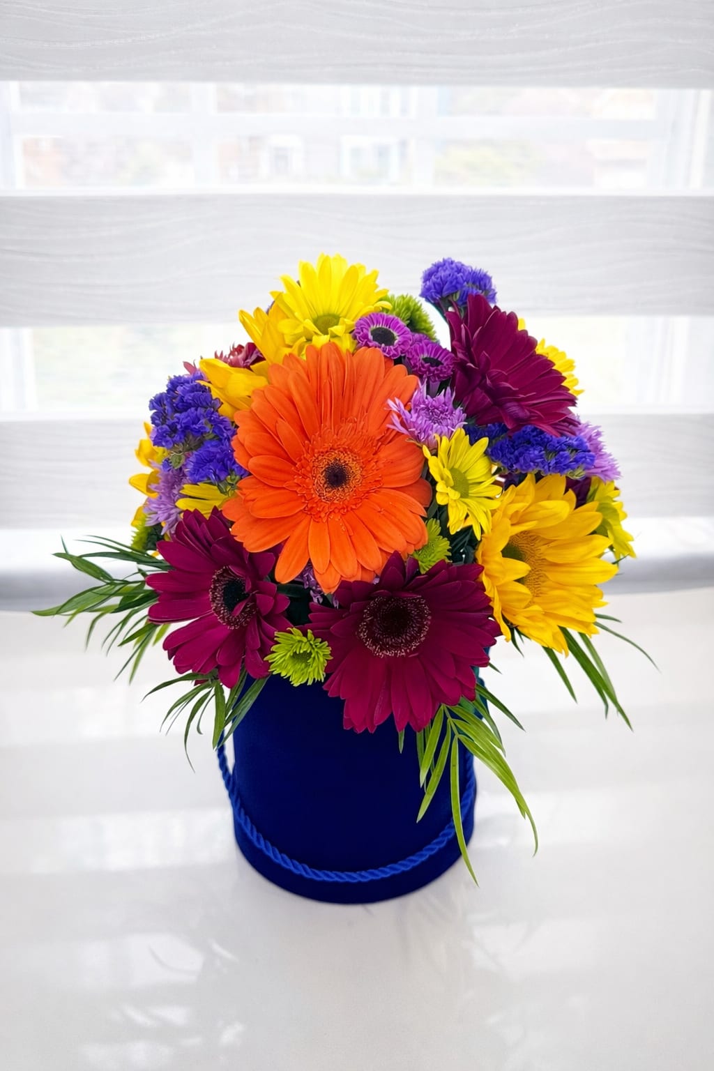Sunburst Gerbera Daisy Hatbox Arrangeme - Bring joy and color to any occasion with our Sunburst Gerbera Daisy Hatbox Arrangement, a lively floral design featuring bright orange, yellow, pink, and purple gerbera daisies accented with fresh greenery.  Designed in a stylish blue hatbox, this cheerful arrangement is perfect for birthdays, celebrations, thank-you gifts, or simply sending happiness. Handcrafted by AZ Flowers &amp; Gifts in Alpharetta, GA, each bouquet is made fresh and delivered with care.   Perfect for: 	•	Birthday surprises 	•	Get well wishes 	•	Thank you gifts 	•	Everyday happiness  We proudly offer same-day flower delivery in Alpharetta, Johns Creek, and nearby areas (30022).