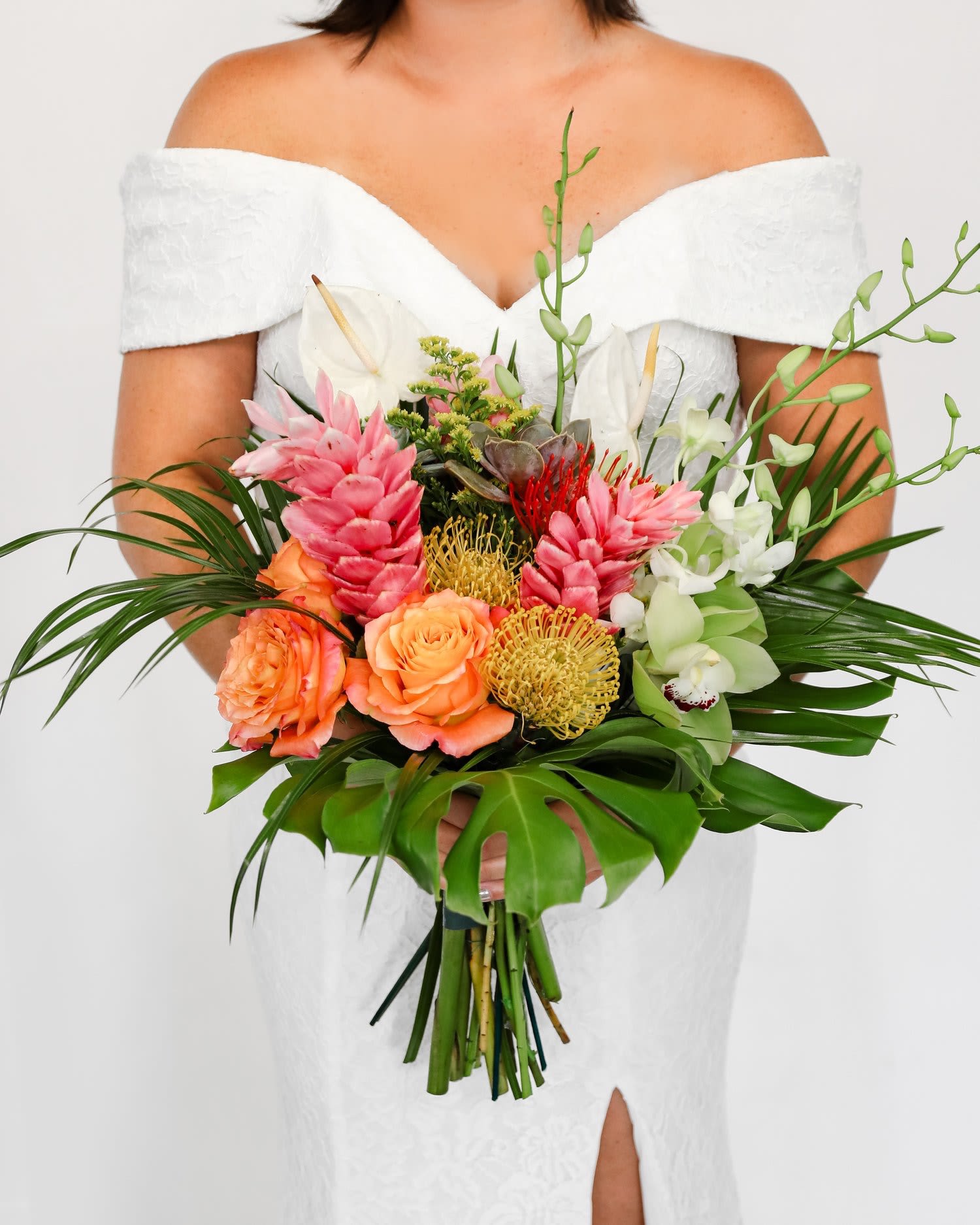 Bride in an off-shoulder white dress holding a large tropical bouquet of pink, orange, green, and white flowers.