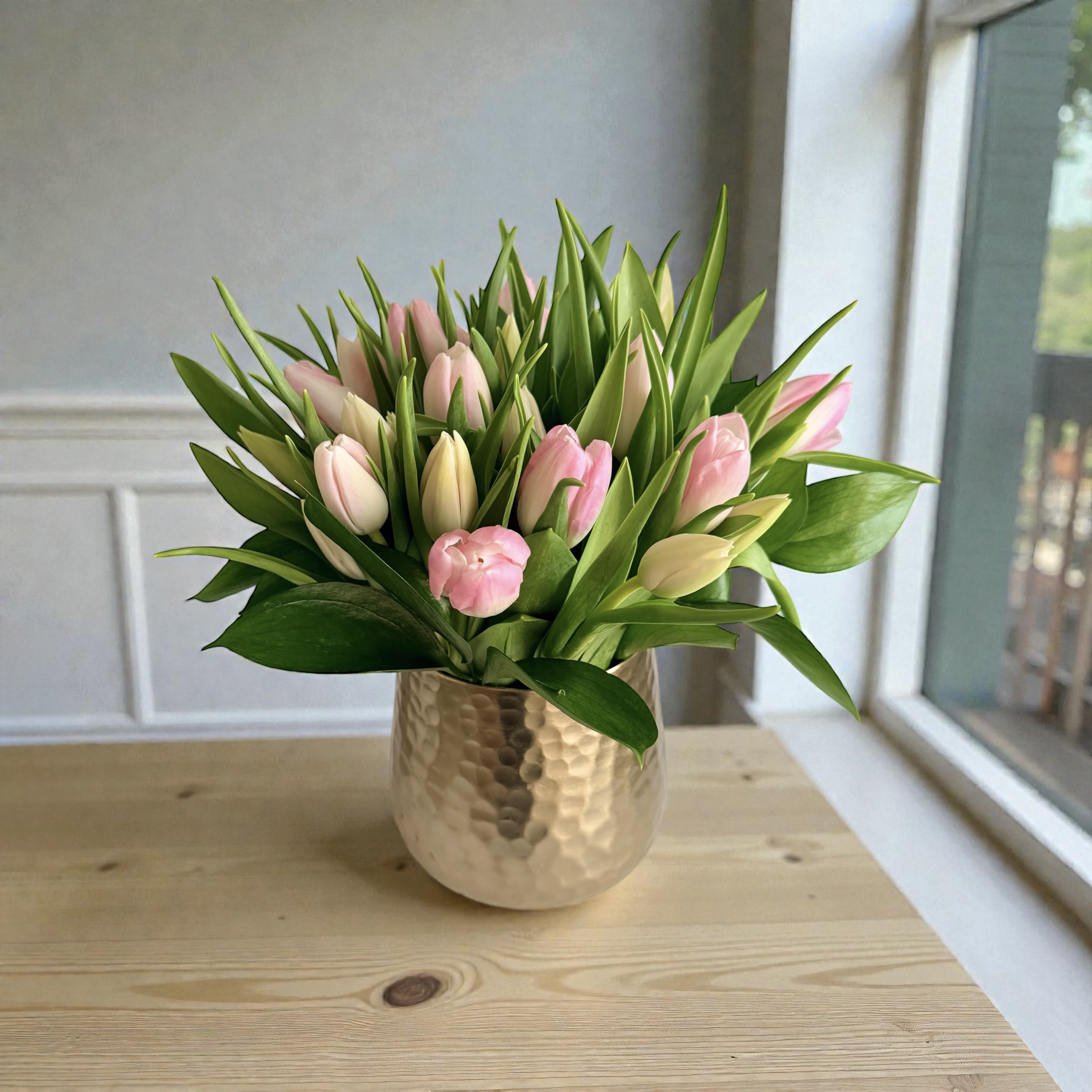 Pink and cream tulips arranged densely in a hammered gold vase on a wooden table by a window