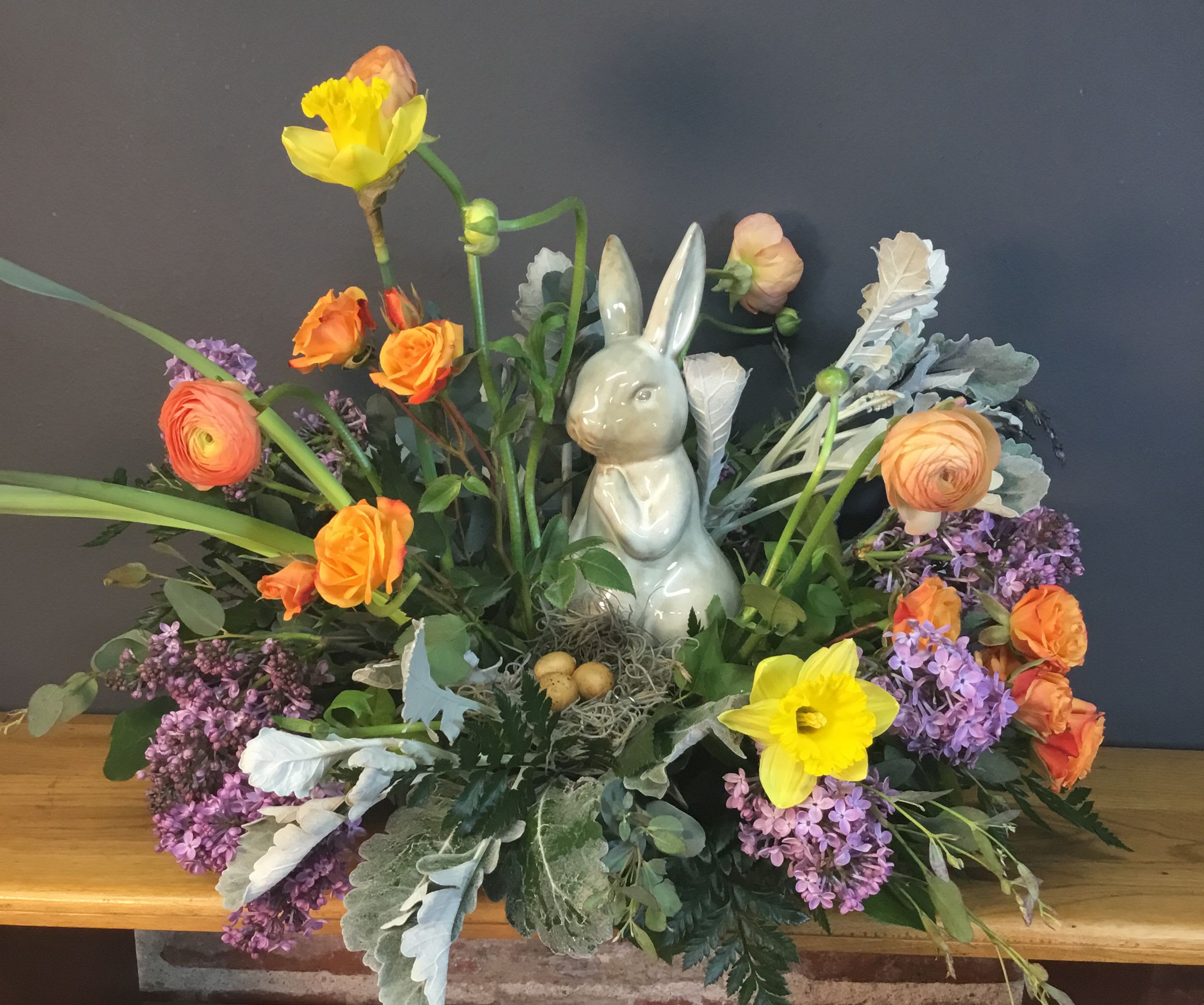 Natural Spring Centerpiece - A playful spring centerpiece that brings the garden indoors-perfect as an Easter table focal or a cheerful seasonal gift. Bright orange roses and ranunculus mingle with soft purple lilac clusters while tall yellow daffodils create upward movement and a sunny focal pop; white accents tie the palette together for a fresh, uplifting look the recipient will notice. This flower arrangement is designed to make someone feel celebrated and joyful-ideal for family gatherings, hostess gifts, or brightening a loved one's home.  The arrangement includes daffodils, roses, ranunculus, and lilac in orange, purple, yellow, and white tones, accented by a white ceramic bunny figurine tucked into the blooms. A small decorative nest with speckled faux eggs and natural textures sits at the base near the bunny to reinforce the seasonal theme. Sized to work as a centerpiece, it arrives ready to display-simply place on a table and refresh water as needed to keep stems healthy for several days.