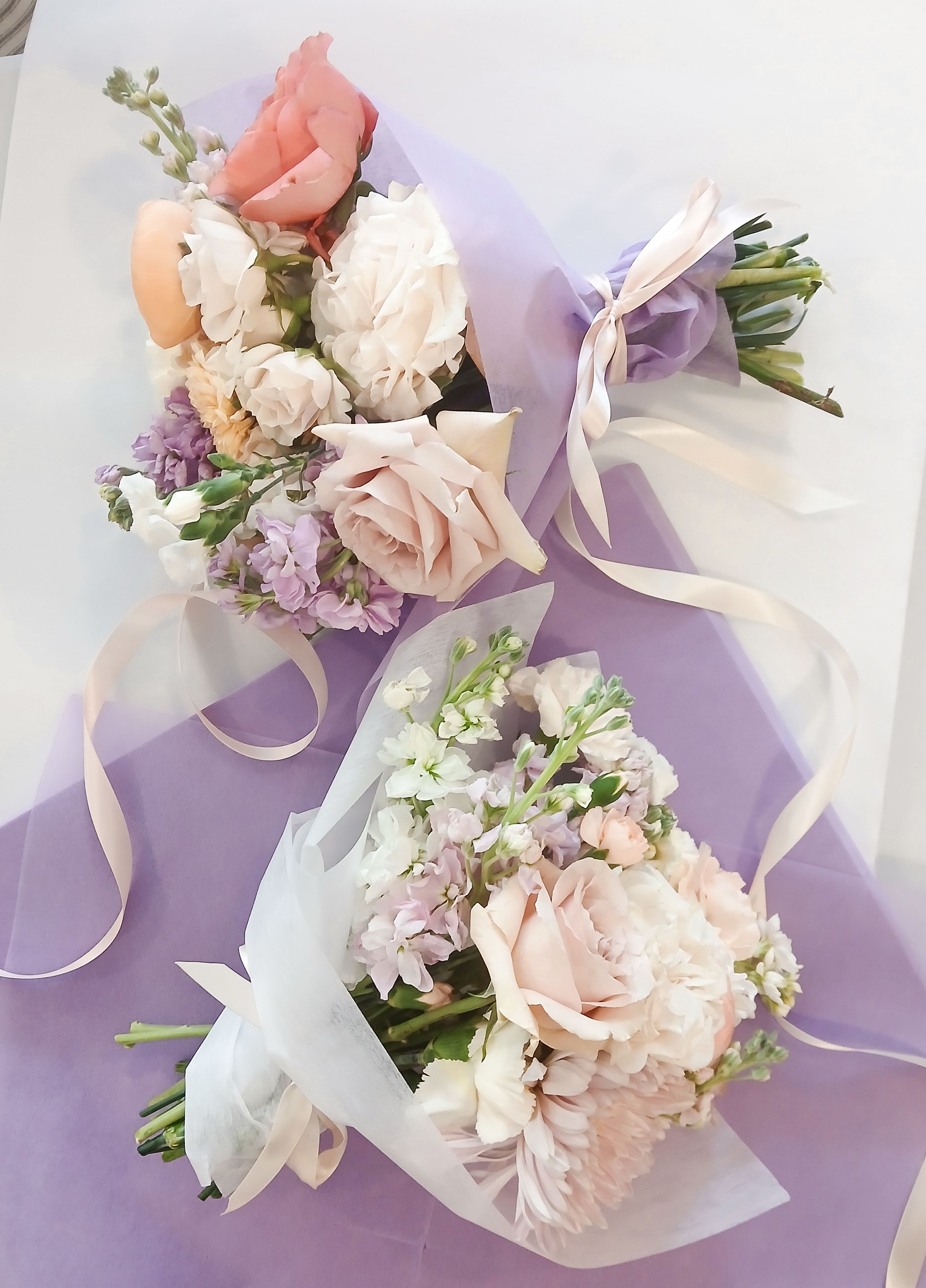 Mother's Day Wrapped Bouquet - Soft pastels and layered texture make this wrapped bouquet feel like a quiet, joyful embrace for the person you're celebrating. A hand-tied flower arrangement for Mother's Day (or any moment you want to mark with tenderness), it combines roses, stock, carnations, and chrysanthemums in shades of blush pink, white, lavender, and peach to create a romantic, airy impression that reads as both elegant and effortlessly kind.  Each bouquet is gathered by hand and finished in pink, lavender or white floral wrap, then tied with a satin ribbon for a refined presentation-two bouquets are shown, each full and textured from the mix of blooms. Expect soft-headed roses, fragrant stock, ruffled carnations, and cushiony chrysanthemums layered to give volume without feeling heavy; the wrap keeps the stems secure and the look light.  Perfect for a mother, mentor, or friend who deserves to feel seen, this arrangement suits birthdays, anniversaries, springtime celebrations, or a thoughtful surprise. Bouquets arrive ready to place in water-trim stems and refresh the vase water every few days to help the blooms last longer.