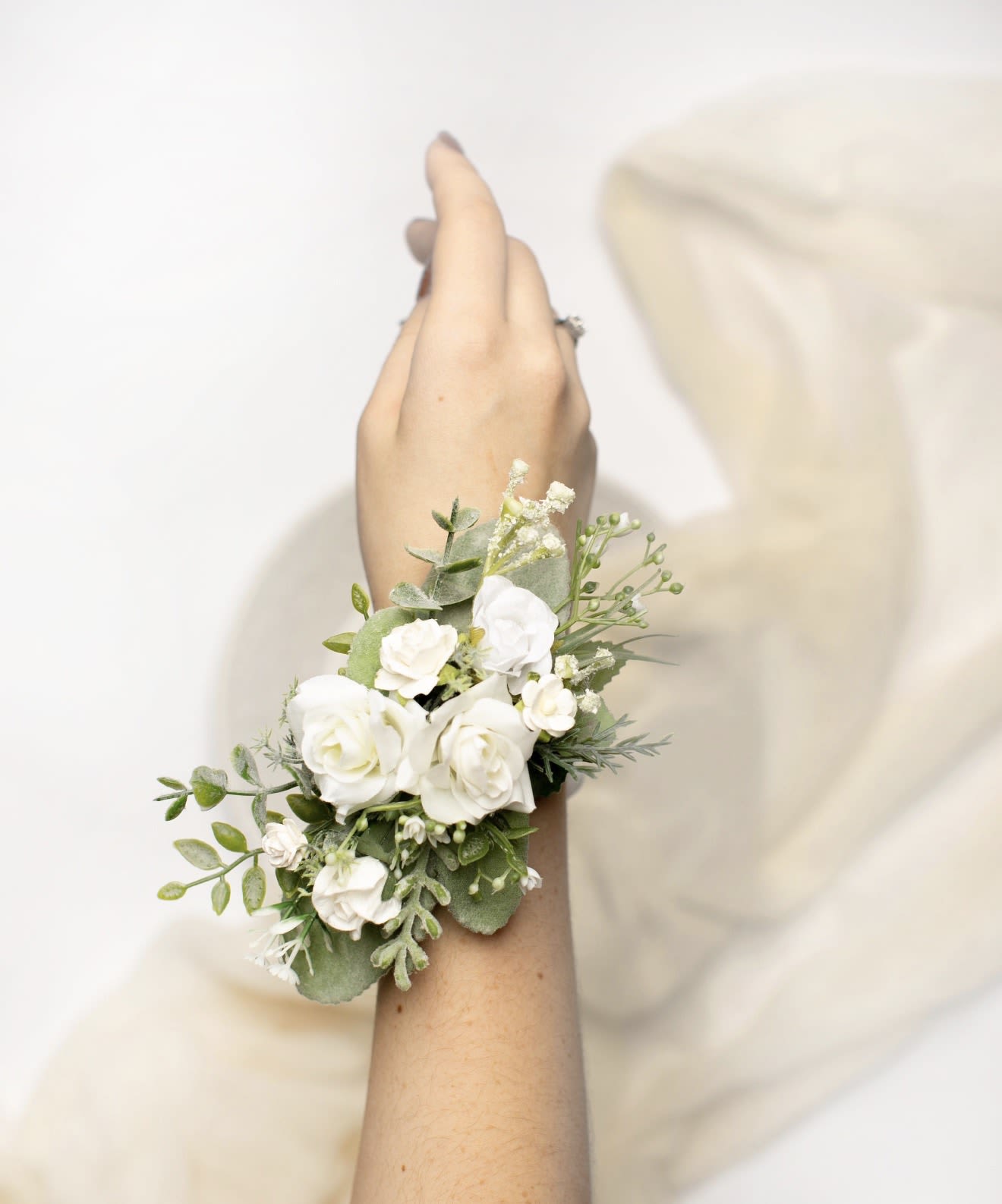 Custom Corsage - A delicate wrist corsage crafted for prom and wedding wear, this Custom Corsage makes the person you're celebrating feel elegantly finished and cherished. The compact cluster design reads classic and bridal with a white-and-green palette that also complements spring occasions like Mother's Day without feeling seasonal-only - it's equally right for formal dances or rehearsal dinners.  Small white spray roses and rounded miniature roses sit amid soft mixed greenery and airy filler, creating a gentle, sculpted silhouette at the wrist. Though sized for subtlety, the addition of textural blooms like tulips, dendrobium orchids, delphinium and dianthus (used as accents when requested) brings depth and a refined contrast between smooth petals and lacy stems. The overall look is clean, fresh, and photograph-ready.  Perfect for the mother-of-the-bride, the prom date, or the bride who prefers understatement, this corsage ties the outfit together while keeping comfort in mind. Ask about exact flower selection or color emphasis when ordering to match bouquets or dresses; we'll ensure proportions suit wrist size and event formality and provide simple care tips so it looks its best through photos and celebrations.