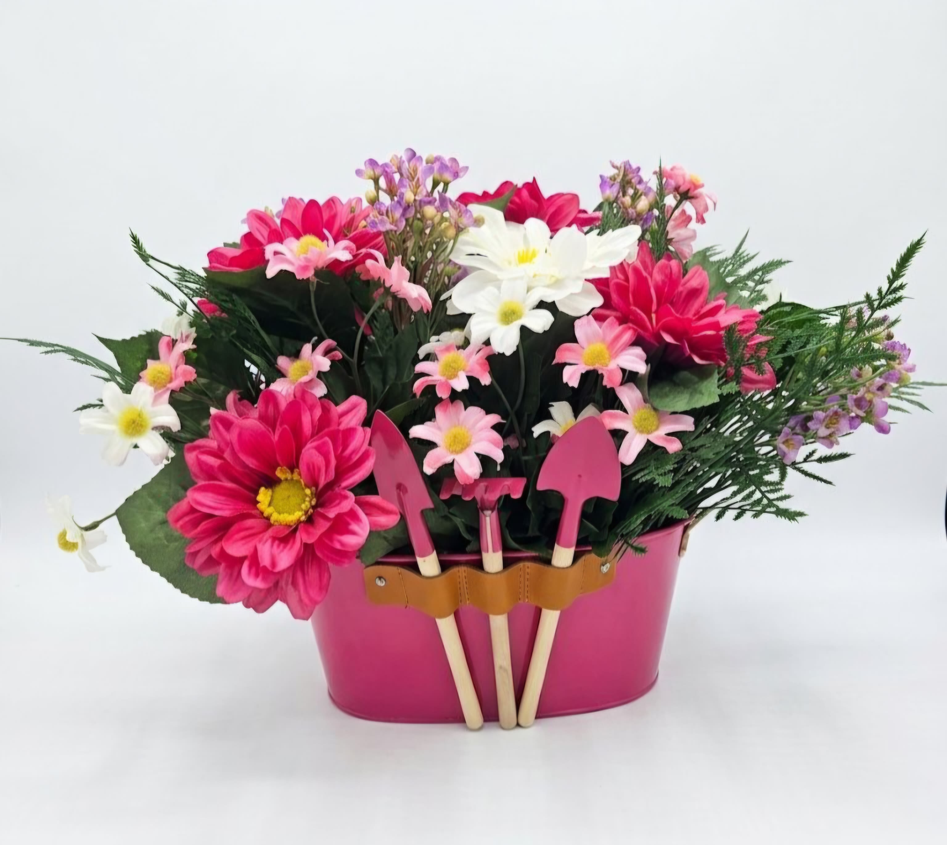 Always in Bloom Pink Gerber Silk Bouquet - A dense cluster of pink and white daisy-like blooms makes this pink gerber silk bouquet feel like a pocket of spring - perfect as a birthday bouquet, a thank-you gift, or a cheerful "just because" surprise. The arrangement reads bright and playful, the vivid pinks and crisp whites softened by airy purple filler flowers so the person you're thinking of will feel instantly uplifted and noticed.  The silk daisies and small purple accents are gathered tightly in a matte pink metal container that doubles as part of the gift. A leather strap across the front holds three small garden trowels in a neat tool holder, giving the piece a charming, garden-inspired personality and extra usefulness as decor for an entryway, sunroom, or office. Colors include pink, white, purple, and green, and the compact design makes it easy to place where it will get the most smiles.  This arrangement is crafted as a casual, everyday present for someone who appreciates bright color and a touch of whimsy - ideal for friends, moms, or coworkers. Because the stems are silk, it stays fresh-looking with almost no care; simply dust occasionally to keep the colors vivid. Order with confidence for a long-lasting bouquet that sends a warm, celebratory message. 11" Tall