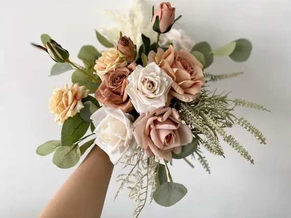 Prom Boquet S2 - Clustered blush, peach, and white roses gather into a compact, rounded prom bouquet that reads like a timeless bridal posy-perfect as a bridesmaid or prom bouquet and for wedding ceremonies wanting soft, romantic florals. The gentle pinks and creamy neutrals create an elegant, understated statement that will make the person you're gifting feel cherished and picture-perfect as they walk down the aisle or onto the dance floor.  Hand-tied in a classic rounded shape, this bouquet pairs roses with delicate airy filler and soft eucalyptus for a loose garden feel and subtle greenery accents. The compact handheld size is designed for comfort and photo-ready looks; colors include pink, white, green, and peach for a versatile palette that complements many gowns and themes. For best appearance, keep stems in water until the moment of use and store in a cool spot away from direct sun prior to the event.