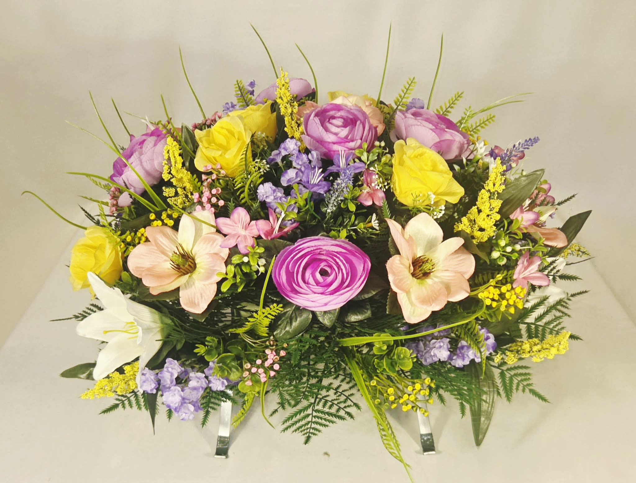 Spring Silk Monument Saddle - A soft pastel cascade of silk roses, lilies, daisies and mixed blooms designed as a memorial saddle sympathy arrangement for Memorial Day, Birthday or in Remembrance. The low, dense composition puts pink, yellow, white and lavender blossoms front and center so the person you're honoring will feel remembered with a gentle, uplifting presence rather than something stark or formal.  The arrangement combines large rose-like blooms and open lily petals with smaller purple and yellow filler flowers for a layered, garden-picked texture that reads full and abundant from every angle. Because it's crafted in silk, the colors stay fresh and the display requires no watering-ideal for a graveside tribute or a table-top memorial. It's the kind of arrangement that quietly comforts and brings a touch of seasonal brightness to the moment.