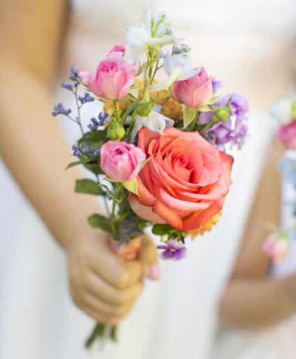 Simplicity bouquet - A single large coral rose anchors this handheld wedding bouquet, surrounded by smaller pink roses and airy white and purple accents for a romantic, garden-fresh bridal bouquet. Perfect for weddings and bridal parties, the compact hand-tied shape keeps the look effortless and the carry comfortable-an intimate choice for a bride who wants pastel charm without fuss.  The bouquet blends roses with mixed seasonal blooms and light-textured fillers to create soft layers of pink, purple, white, and orange-coral. Varied bloom sizes add gentle movement and a delicate silhouette; stems are gathered in a neat, informal wrap so the arrangement reads cohesive from every angle.  Made to suit ceremonies, photos, and reception moments, this bouquet makes the person carrying it feel tenderly celebrated and seen. Hand-carry dimensions keep it easy to hold throughout vows and portraits; treat with fresh water until ceremony time and store cool to preserve petals and fragrance.