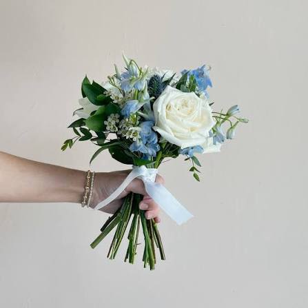 Just Right Bouquet - A large white rose takes center stage in this hand-tied wedding bouquet, its creamy petals balanced by soft blue blooms and delicate white filler for a fresh, romantic bridal look. Designed as a wedding bouquet, it creates an airy, elegant impression that will make the person carrying it feel gracefully seen on their ceremony day.  The compact, hand-held arrangement pairs loose greenery and long stems gathered beneath a pale ribbon for a natural, easy-to-carry silhouette. Textural contrasts between the smooth rose, soft blue florals, and wispy filler give visual depth while the cool white, blue, and green palette keeps the overall feel refined and serene.  Perfect for a bride, bridesmaid, or as a ceremony piece, this bouquet suits classic, coastal, or garden weddings and reads beautifully in photographs. Stems arrive gathered and ribboned for immediate use-light misting and fresh water in a vase before the event will keep blooms at their best.