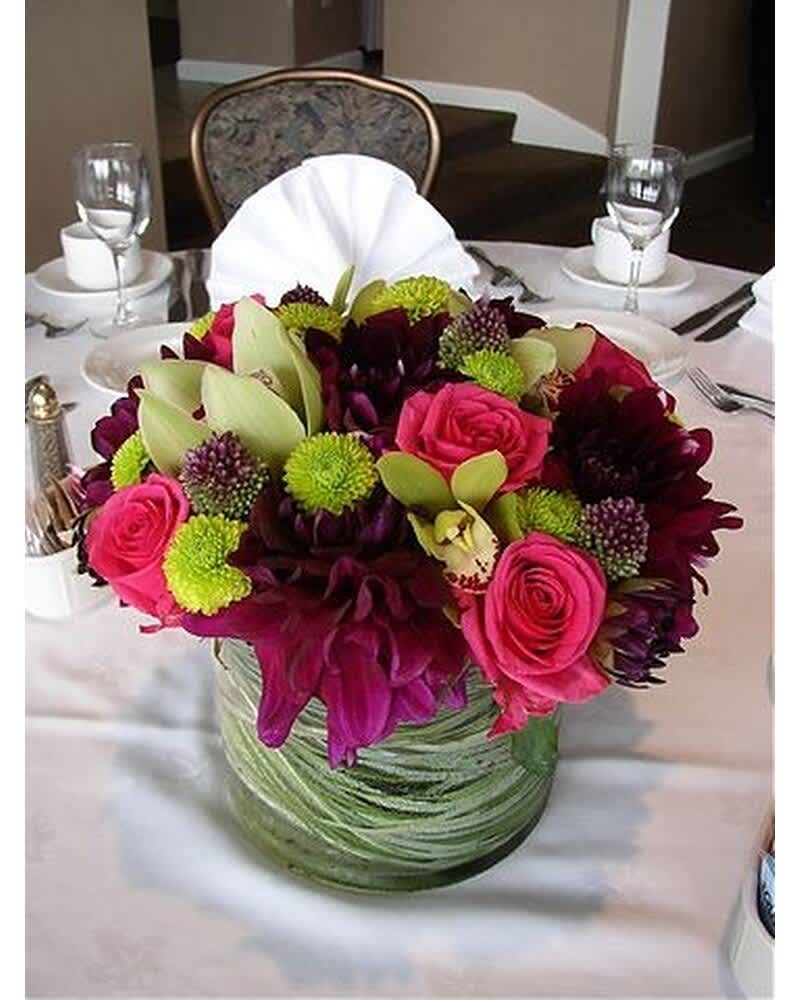 Low centerpiece of pink roses, dark purple blooms, green mums and orchids in a glass cylinder vase