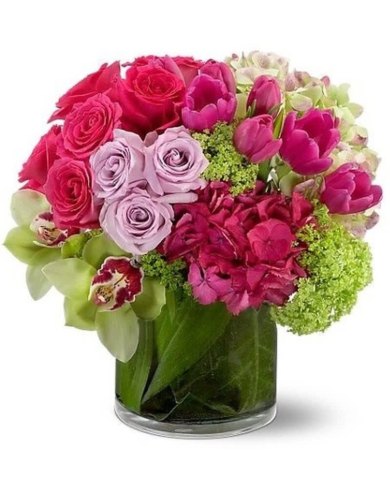 Compact arrangement of pink and lavender roses, tulips, hydrangeas and green orchids in a clear glass vase