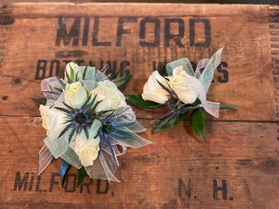 Two white floral boutonnieres with blue accents on a wooden surface