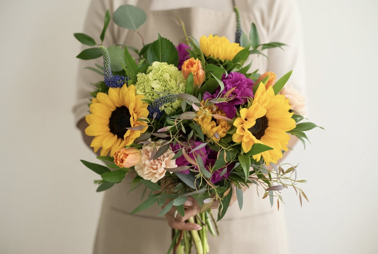 Hand-tied bouquet with bright yellow sunflowers, purple blooms, and mixed flowers held by a person in beige.