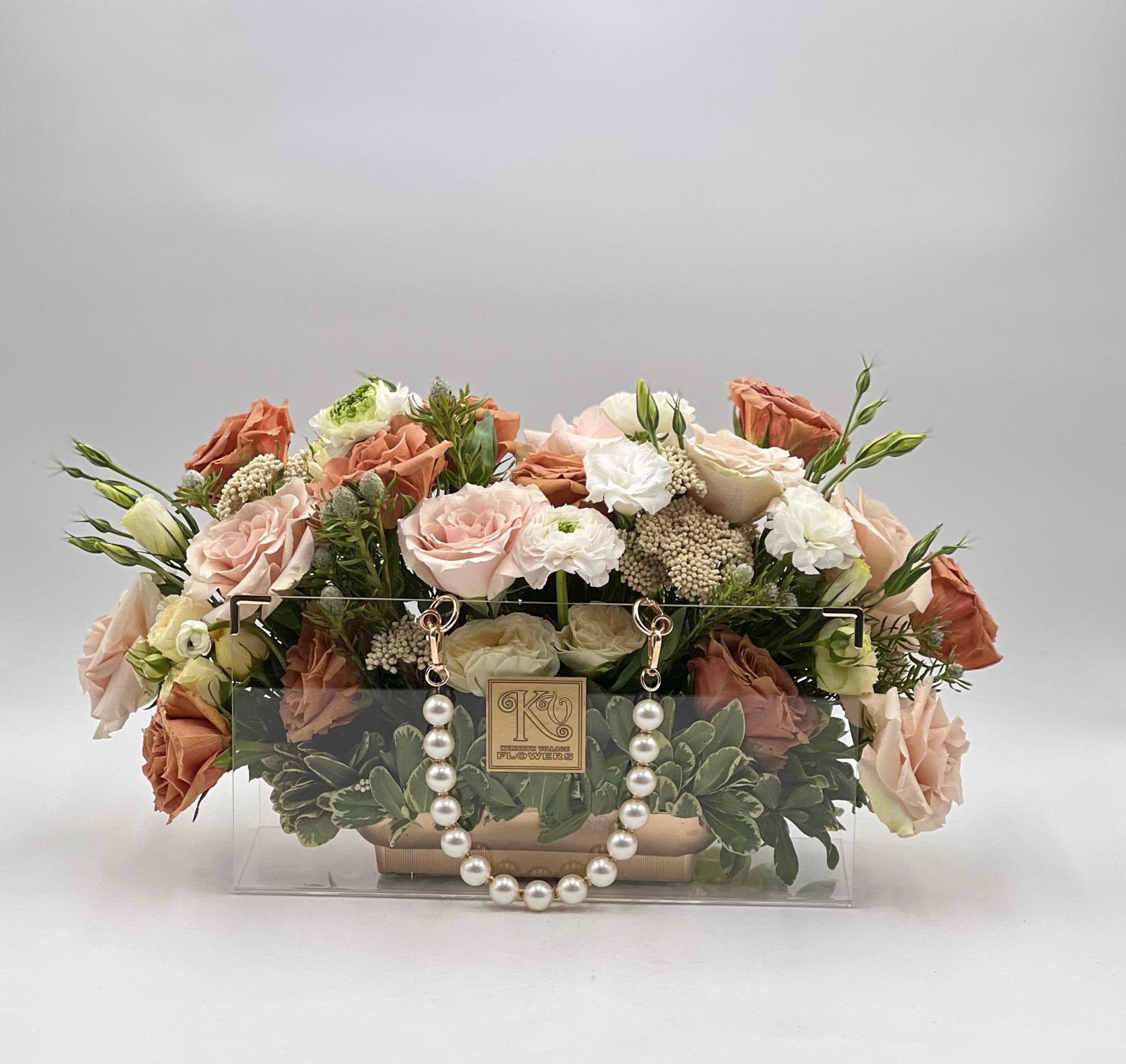 Pearl Blush Luxe Handbag - A refined blend of soft blush, ivory, and warm peach tones, this elegant arrangement is designed in a signature clear handbag-style box adorned with delicate pearl accents. Featuring premium roses, textured blooms, and lush greenery, it creates a romantic and sophisticated look with a modern twist. Perfect for any occasion, this design offers a timeless expression of beauty and grace, making it an unforgettable gift or centerpiece.