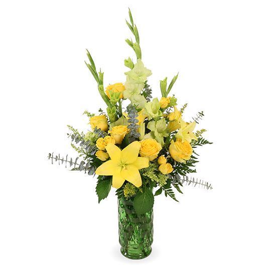 Tribute Dawn - Sunlit yellow lilies and clustered yellow roses give this sympathy-style flower arrangement a warm, uplifting presence that brightens any room. As a cheerful sympathy or get well bouquet, Tribute Dawn blends vertical pale-green snapdragon spikes and mixed foliage to create an airy, graceful silhouette that feels both comforting and hopeful.  The flowers are arranged with textured filler accents for lively texture and gentle movement, all presented in a textured green glass vase that reinforces the springlike yellow, green, and white palette. The tall, vertical design makes a fresh statement on a side table, entryway, or hospital room and is the kind of arrangement that helps the person you're thinking of feel genuinely comforted and seen.