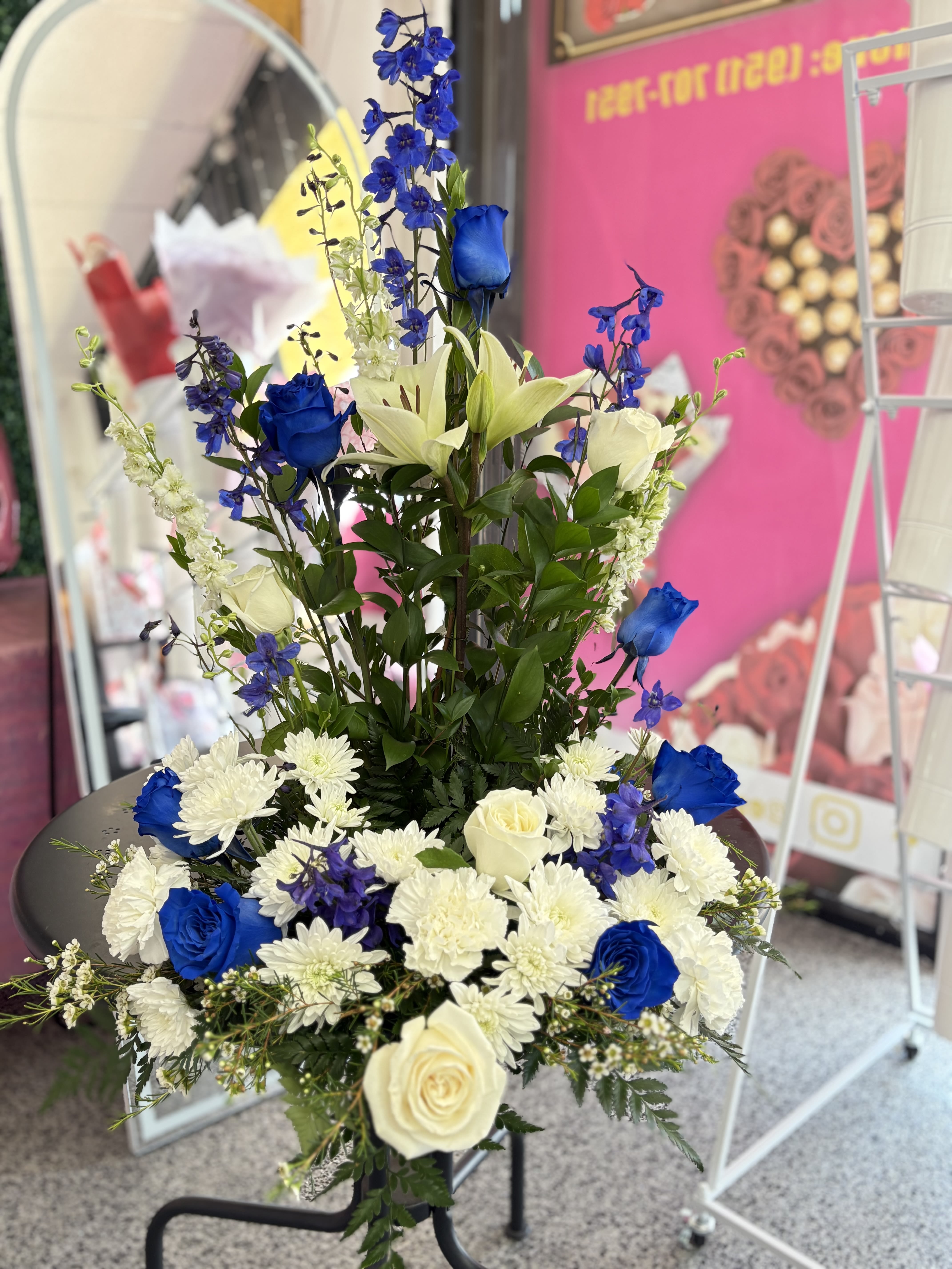 Blue and White urn arrangement  - Vivid blue roses rise beside pure white lilies and roses in this blue and white urn arrangement, a tall flower arrangement that makes an immediate, refined impression. Designed for occasions ranging from a dignified sympathy tribute to a formal celebration, the upright composition and crisp blue-and-white contrast ensure the person you're honoring feels seen and respected from across the room.  Layered and structured, the design features dense florals at the base that taper into a narrower vertical center framed by spiky blue delphinium and clusters of white daisies for texture and movement. Hues of white, blue, and fresh green foliage create a cool, elegant palette; the arrangement is presented on a floral stand so it's intended for display where height and presence matter.  Ideal for memorial services, dedications, or any moment that calls for a stately, composed gesture, this piece reads as both respectful and celebratory depending on the setting. Because it's designed to be viewed from a distance, allow its strong vertical lines and color contrast to speak for you when you want the recipient to feel honored and comforted.