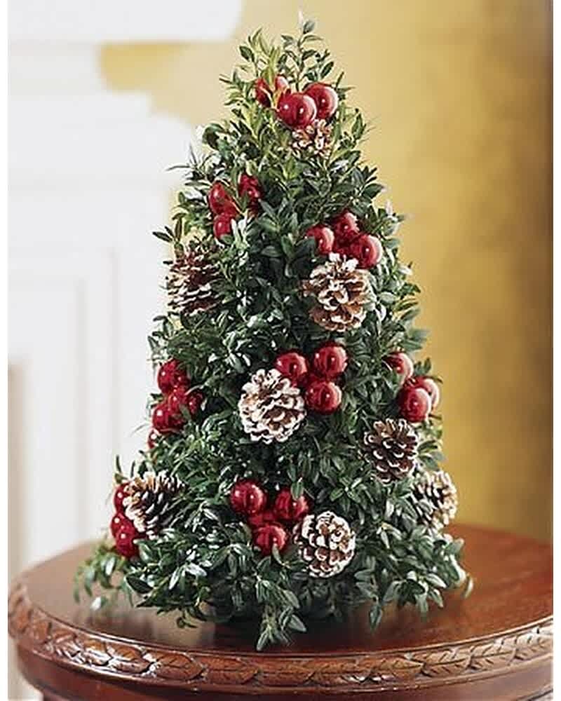 Tabletop evergreen tree decorated with red berries and frosted pine cones on a wooden pedestal table.