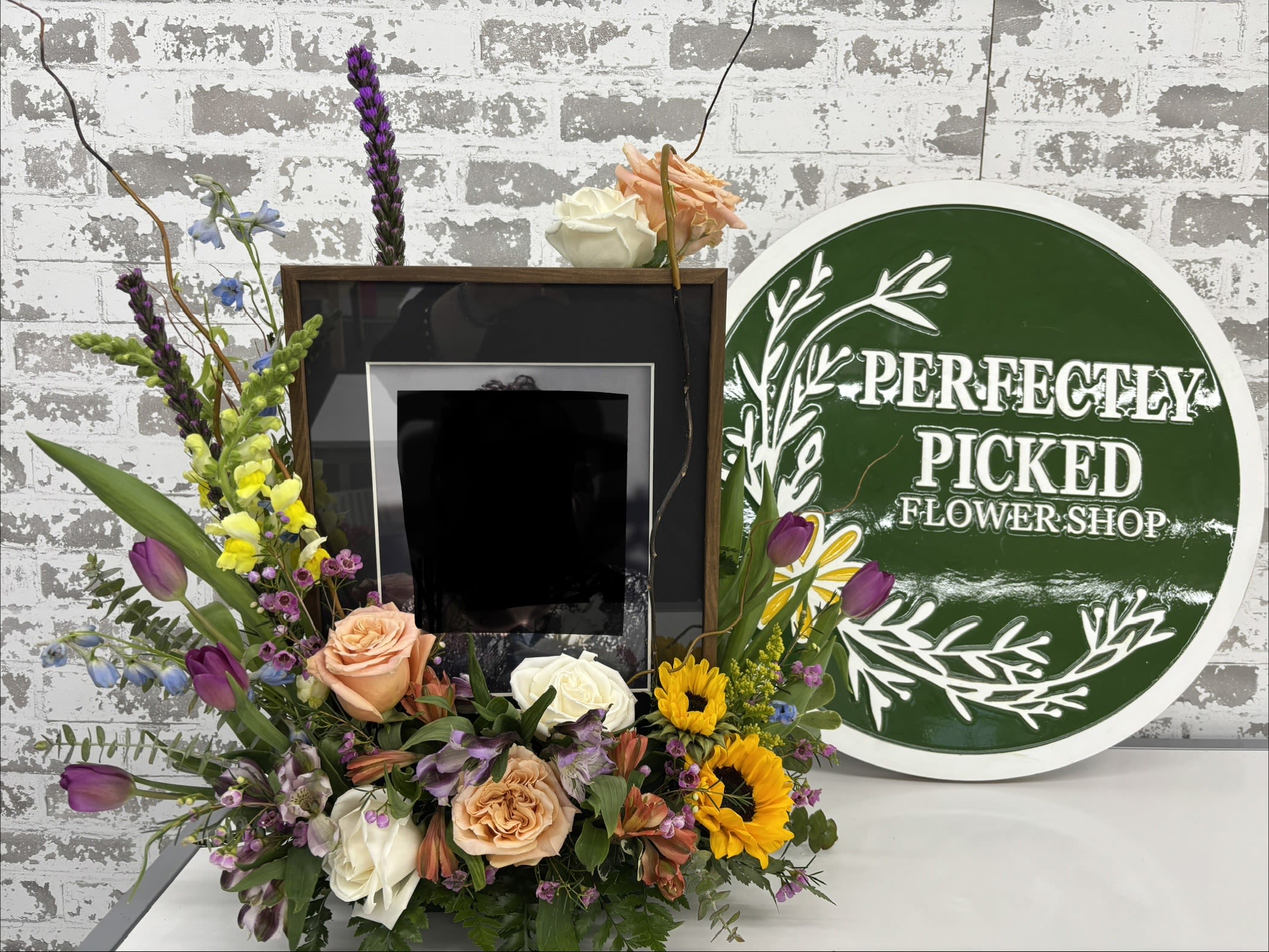 Photo Tribute Memorial  - Surrounding the frame is a lush mix of fresh flowers. The vertical elements, and natural branches, give the arrangement height and a graceful, uplifting field, while the greenery provides fullness and a natural, garden like look.