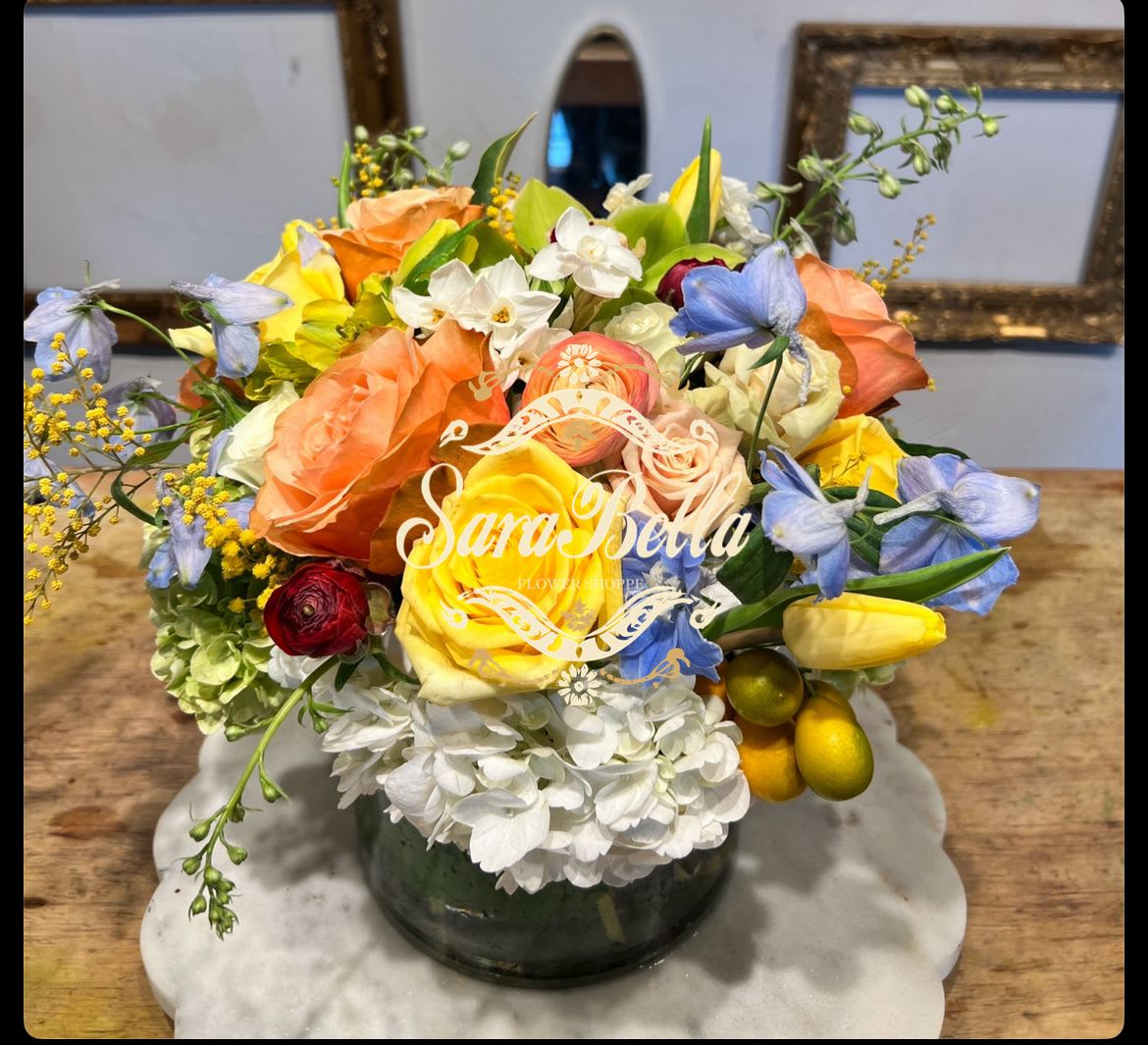 CHEERFUL AND FRESH - A cheerful,garden-inspired arrangement featuring vibrant yellow and peach roses, soft blue accents, and delicate white blooms... 