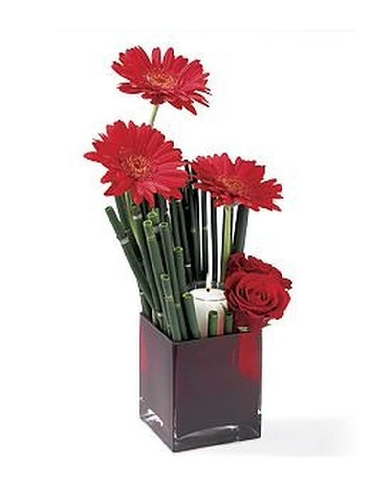 Modern arrangement of red gerbera daisies and roses in a red cube vase with a white candle.