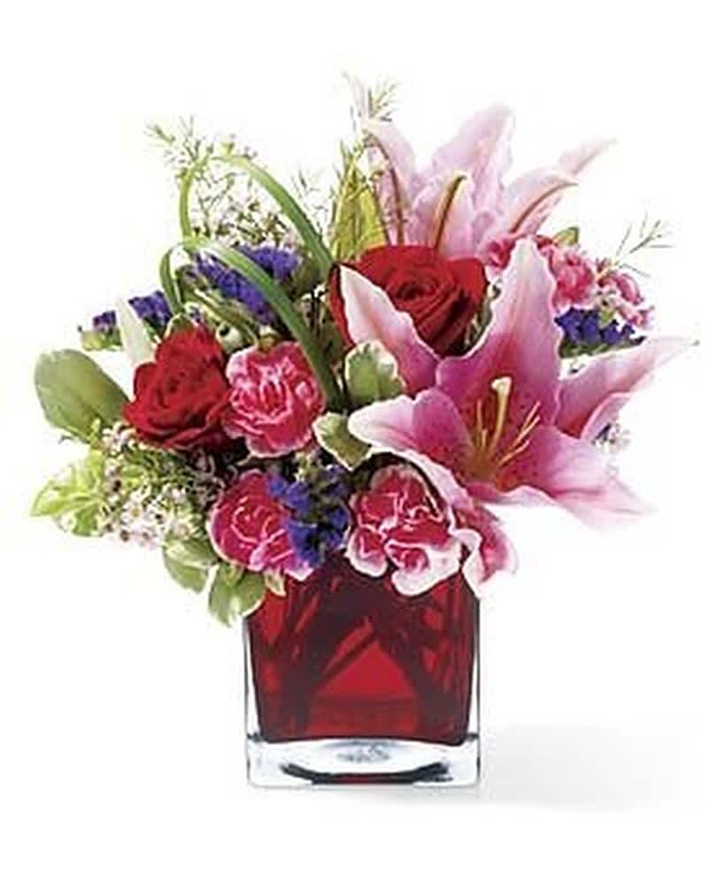Pink lilies, red roses, and pink carnations arranged in a red glass cube vase
