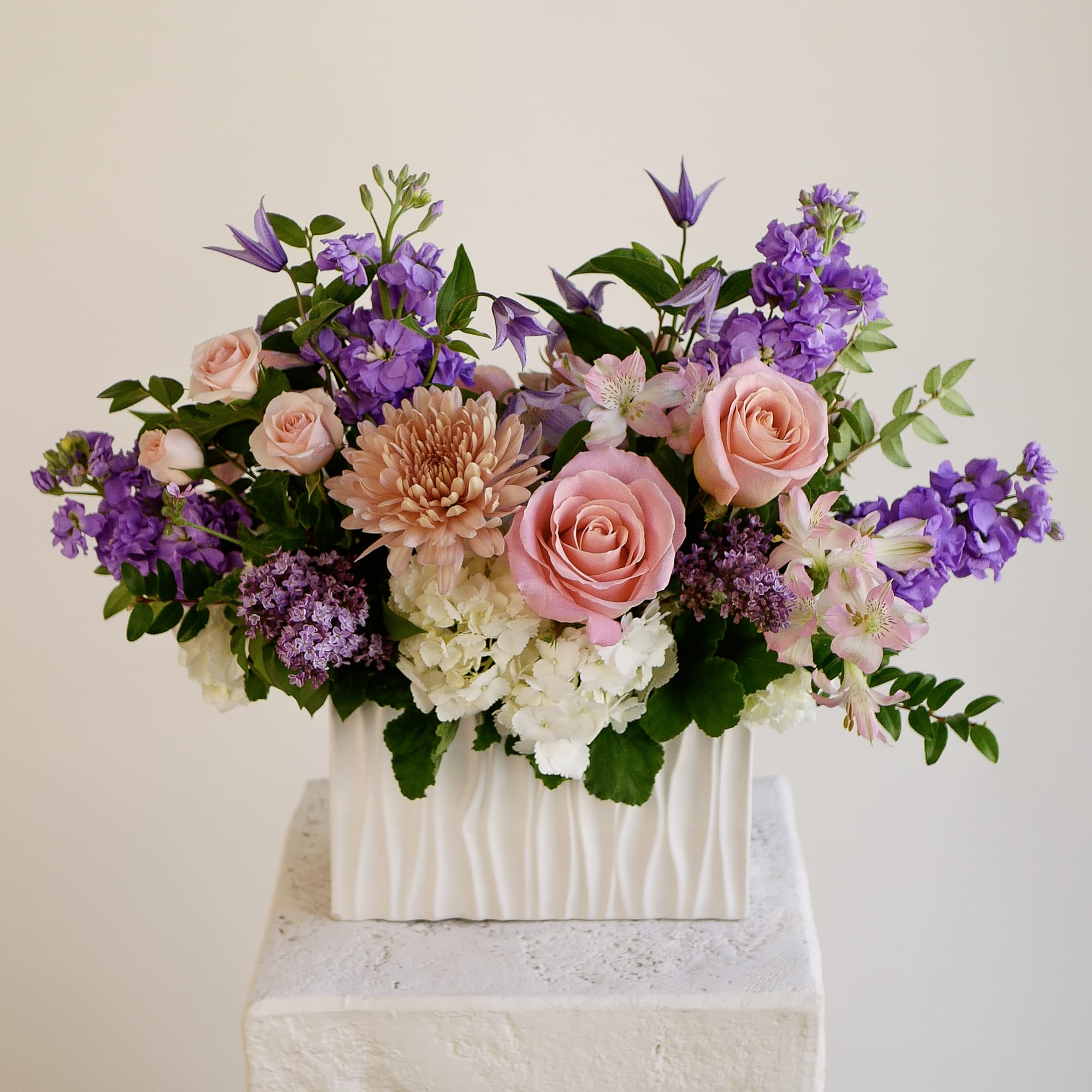 Lilac Love - Soft pink roses and vibrant purple stock create a lovely palette, accented by peach chrysanthemum, white hydrangea, and delicate pink alstroemeria. Purple clematis adds unexpected elegance while fresh greenery provides natural texture throughout. Arranged in a wide ceramic vase, the piece sits approximately 21 inches wide and 16 inches tall—perfect for dining tables, kitchen counters, or entryways. Every stem is hand-selected by our designers for color harmony and long vase life. Local same-day delivery is available across West LA neighborhoods, ensuring the flowers arrive at peak freshness.