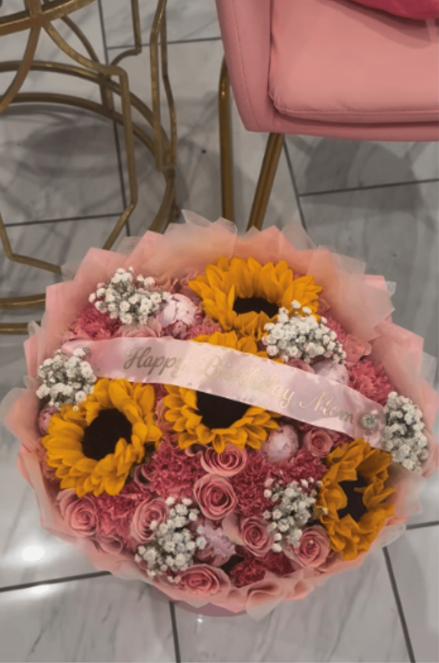 Pink Delight - This flower arrangement features tightly packed sunflowers, pale pink roses, ruffled pink carnations (and similar soft pink blooms), plus clusters of small white filler flowers arranged and wrapped in pale pink tissue and water proof paper . A custom message printed light-pink ribbon that reads "Happy Birthday Mom," and a decorative wrap frames the flowers for a polished, gift-ready presentation.