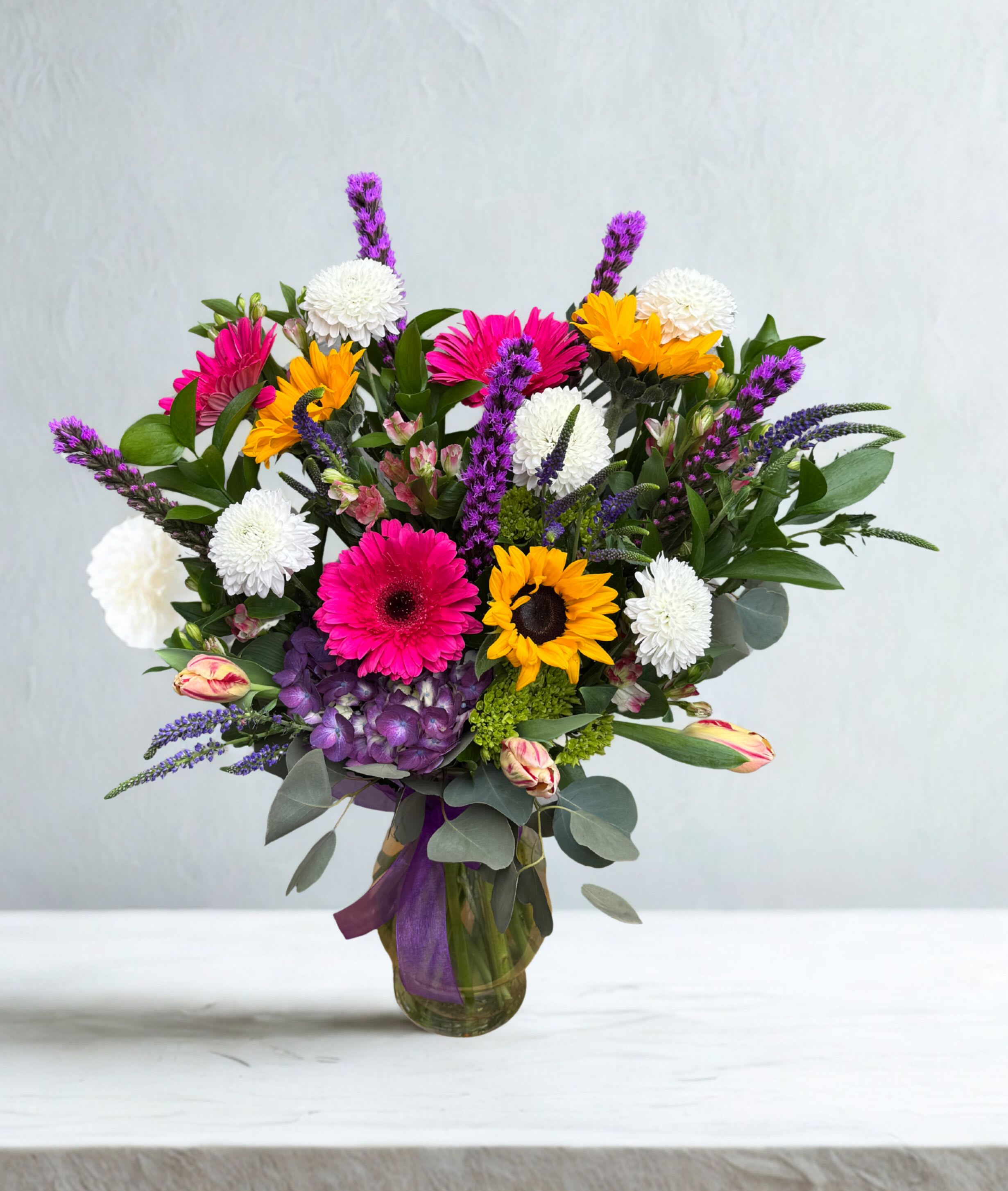 Sunshine &amp; Smiles - Bright, cheerful, and full of personality, this vibrant arrangement is designed to make a statement. Featuring bold sunflowers, hot pink gerbera daisies, crisp white blooms, and rich purple accents, it brings together a playful mix of color and texture. Lush greenery and delicate filler flowers add depth and movement, all beautifully arranged in a clear glass vase with a soft ribbon finish.