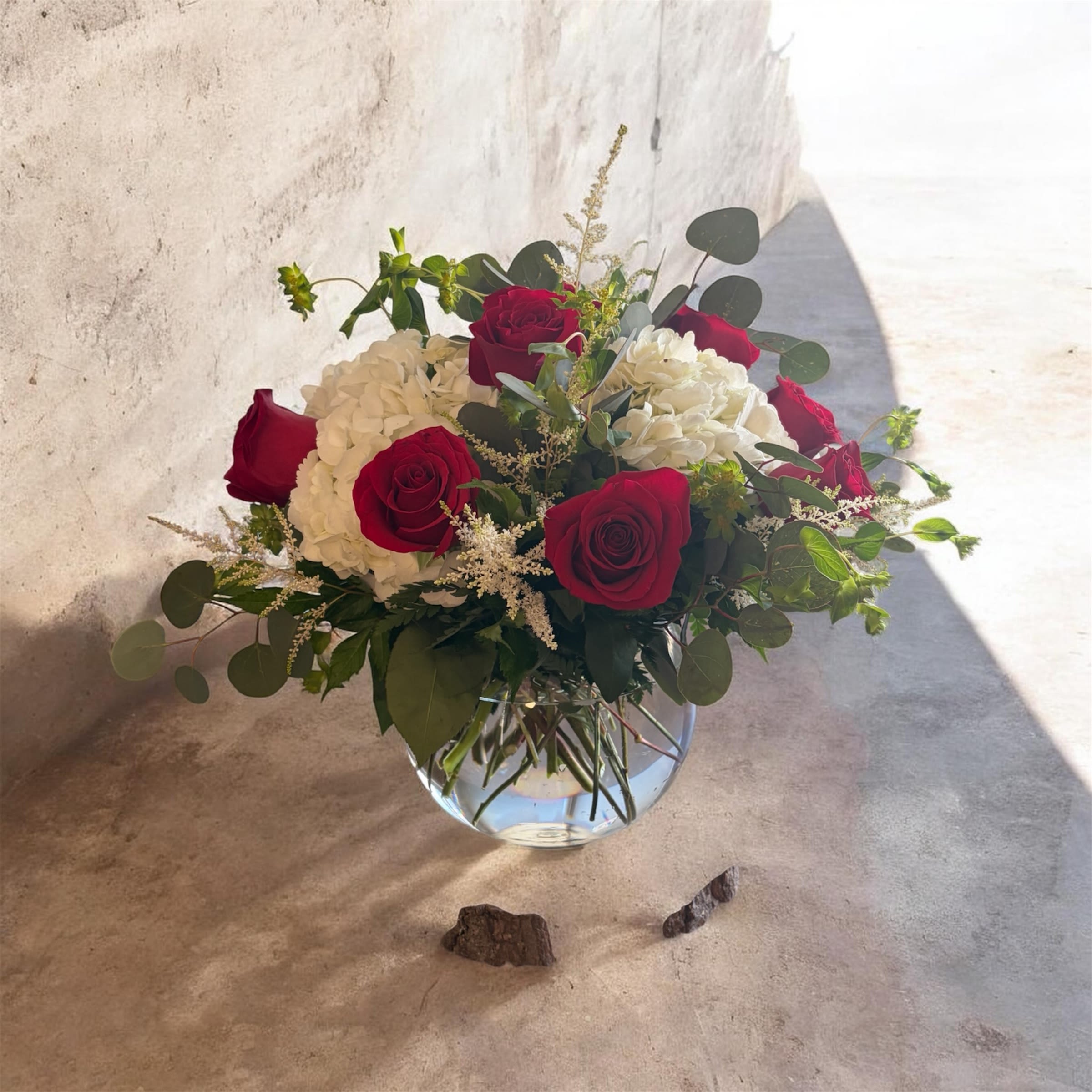 Rose bowl - Deep red roses paired with billowy white hydrangeas form a striking rose bowl arrangement that reads romantic and refined - perfect for an anniversary bouquet, a love-themed gift, or a respectful sympathy arrangement. The bold red-and-white contrast delivers immediate visual impact so the person you're thinking of will feel noticed and cherished the moment the arrangement arrives.  Set in a rounded clear glass vase, the medium-sized design combines classic roses and large hydrangea blooms with airy greenery and delicate filler stems for a loose, garden-style silhouette. The greenery extends outward for gentle movement while the compact bowl shape keeps the composition feeling full and fresh. Colors are vivid red, crisp white, and lush green, making this a timeless choice that pairs easily with many settings.  Ideal for celebrating a milestone, expressing heartfelt sympathy, or simply saying "I love you," this arrangement arrives ready to display in its clear glass vase. For best longevity, refresh the water and trim stems every few days to keep the blooms looking their best.