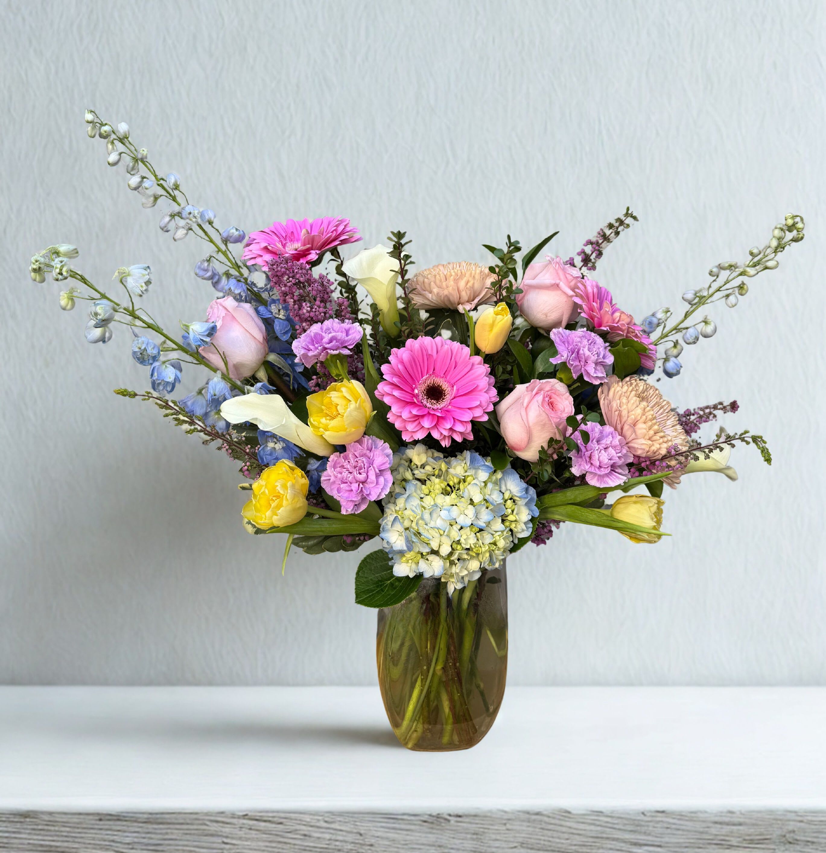 Spring Meadow - Soft, airy, and full of charm, this garden-inspired arrangement blends delicate pastels with a fresh, natural feel. Featuring blush roses, cheerful gerbera daisies, hydrangea, tulips, and seasonal accents in shades of pink, yellow, lavender, and blue, it creates a beautifully balanced and uplifting design.