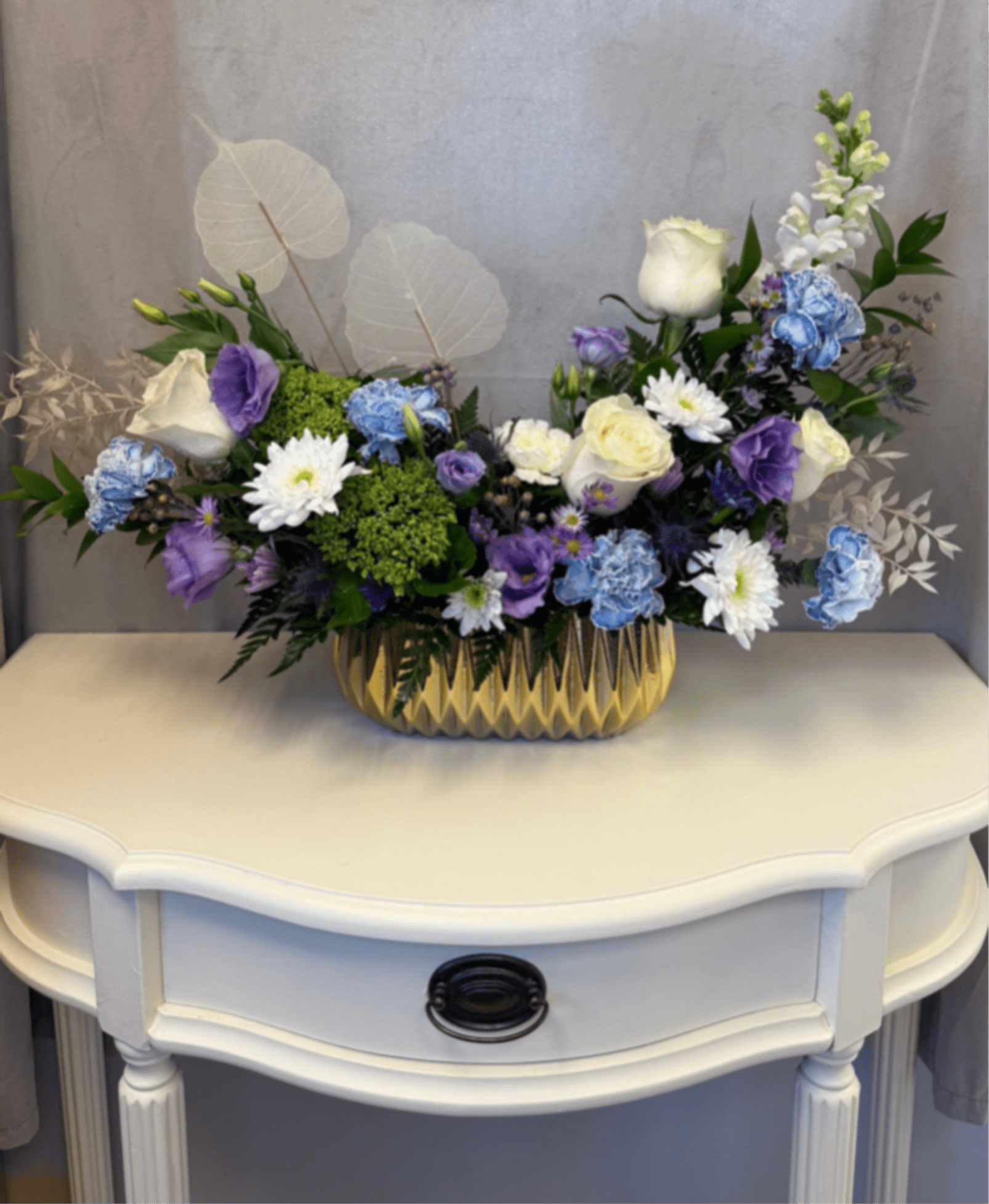 Lavender Mist - Lavender Mist is a soft floral arrangement that captures the beauty of a dreamy garden in bloom. Blending delicate shades of lavender,  blue, and white, this design features a curated mix of roses, lisianthus, carnations, green hydrangeas and seasonal accents for a naturally elegant look.  Airy textures and preserved skeleton leaves add a modern, ethereal touch, while the luxurious vessel elevates the entire piece Lavender Mist feels both effortless and sophisticated.  Perfect for thoughtful gifting, intimate celebrations, sympathy gestures, or as a statement centerpiece, this arrangement brings a sense of calm beauty and timeless style to any space. 