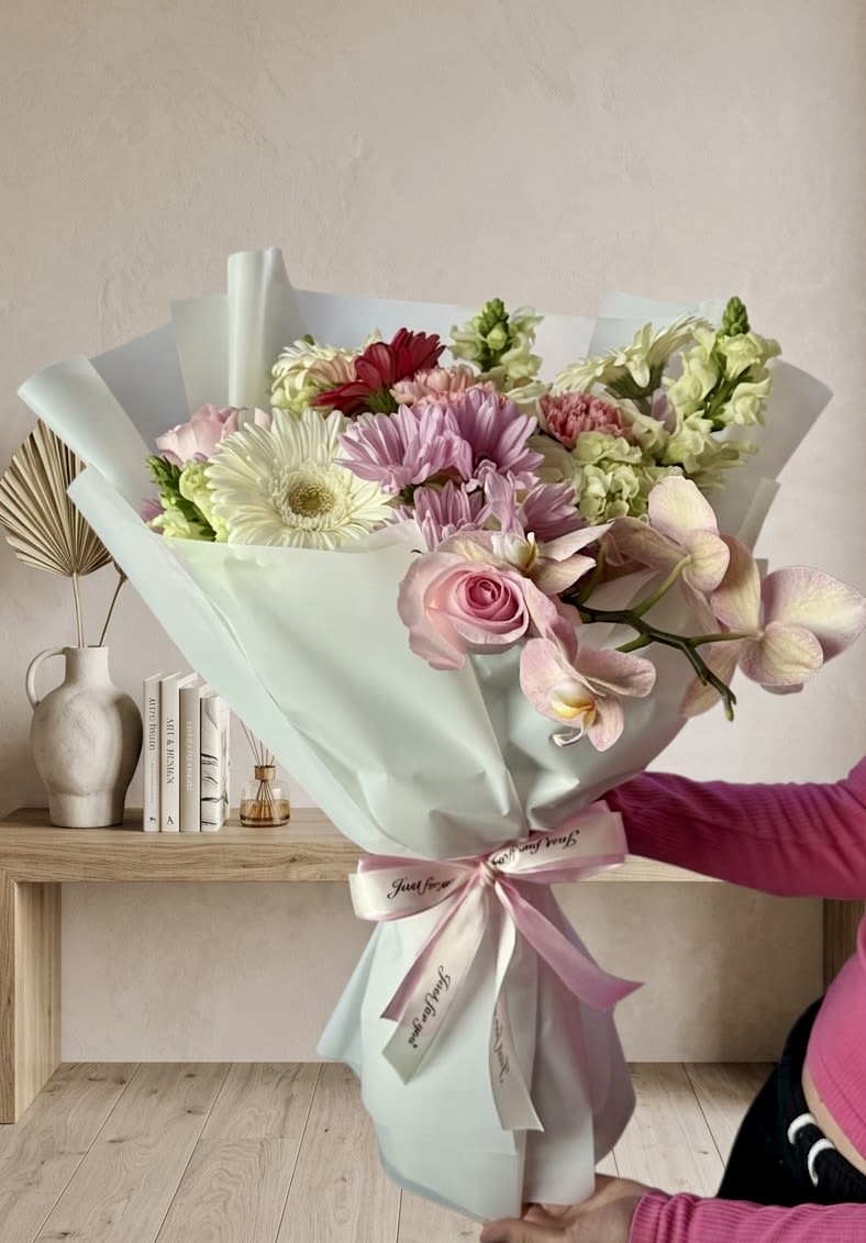 Golden Blooms - Soft pink gerbera daisies and creamy roses mingle with lavender orchids and white chrysanthemums in a romantic, cheerful arrangement perfect as a birthday bouquet, anniversary gift, or a thoughtful way to say you're thinking of someone. The mix of large daisy-like blooms and clustered carnations creates a full, rounded silhouette that reads both celebratory and intimate-the single deeper red accent bloom gives just the right touch of contrast to draw the eye.  Wrapped in pale floral paper and tied with a satin ribbon, the arrangement arrives gift-ready with trailing orchid stems that lend delicate movement. Colors include pink, white, purple and mixed tones; materials include floral wrap and a satin ribbon for a refined finish. Sized to feel generous without overwhelming a tabletop, this bouquet makes the recipient feel seen and celebrated-order with same-day delivery where offered and keep blooms in fresh water, away from direct heat, to extend their vase life.