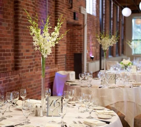 Tall white floral centerpieces on round banquet tables in a brick-walled room