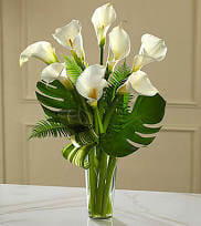 White calla lilies arranged in a clear glass vase with large tropical leaves
