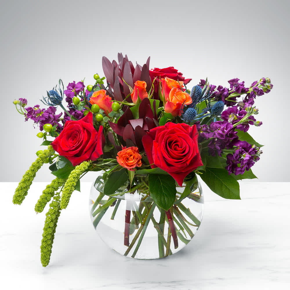 Mixed bouquet of red roses, orange blooms, and purple flowers in a clear glass vase