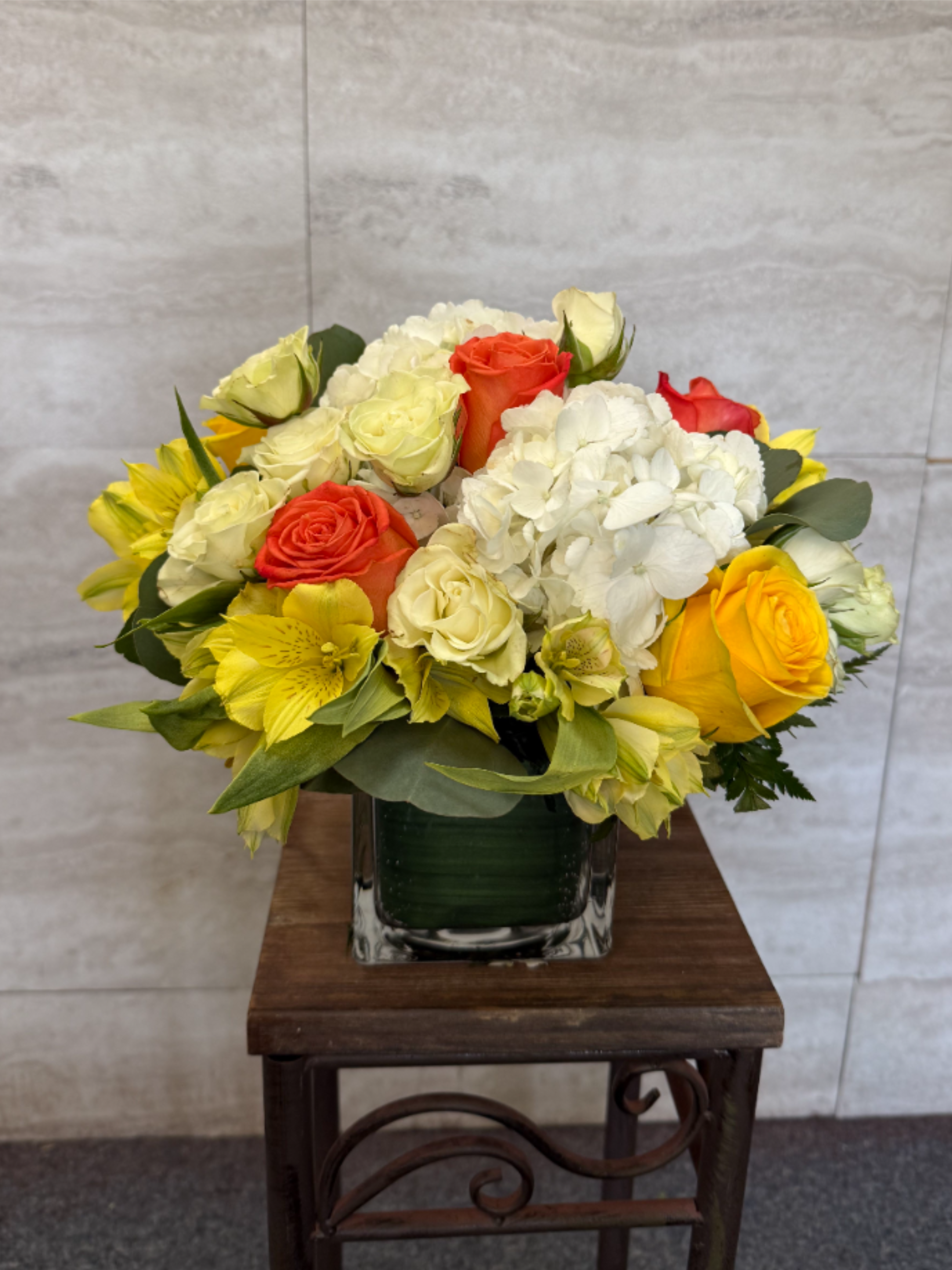 Compact arrangement of yellow, white, and orange flowers in a clear glass cube vase.