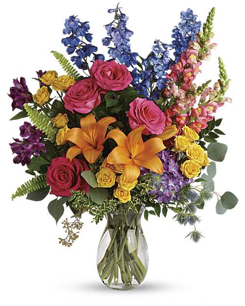 Tall mixed bouquet of pink roses, orange lilies, and bright assorted blooms in a clear glass vase.