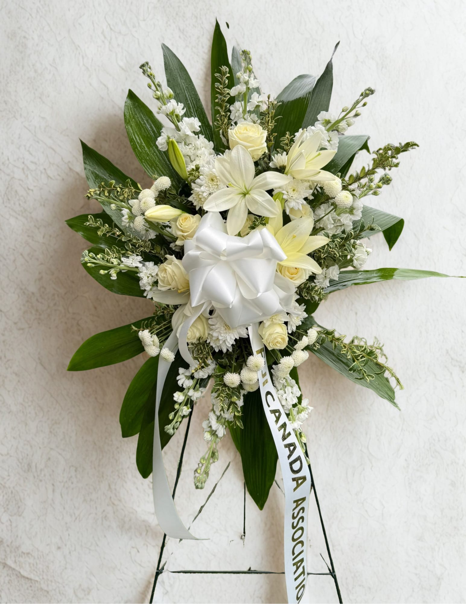 Eternal Lillies - A large white standing spray designed for sympathy or memorial services, this arrangement centers on layered lilies and roses to convey a solemn, comforting presence. The white and green color palette and formal structure make it an appropriate choice for funerals, memorials, or to send condolences-the person you're honoring will feel respected and remembered.  The spray features prominent white lilies, classic white roses, white chrysanthemums, and delicate white filler blooms arranged against broad dark green leaves that fan outward for a structured, ceremonial silhouette. A large white bow anchors the composition at the center and a printed ribbon hangs below; the arrangement is mounted on a standing easel for prominent display.  Choose this arrangement to express sympathy or to honor a life with understated elegance-it's the kind of floral tribute that helps mourners feel seen and supported. Sized as a large standing spray, it arrives ready to place on the easel; for best longevity, keep the spray in a cool, sheltered location until the service.