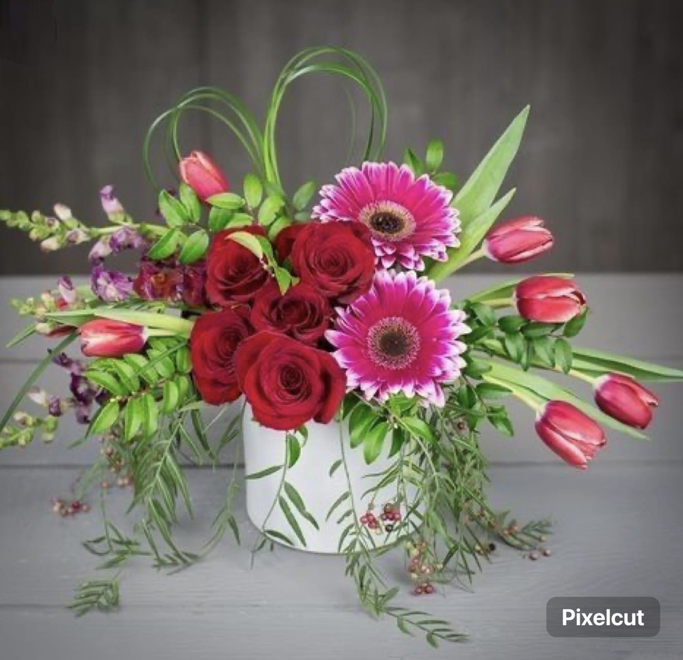 Red roses and pink gerbera daisies in a white vase with trailing greenery
