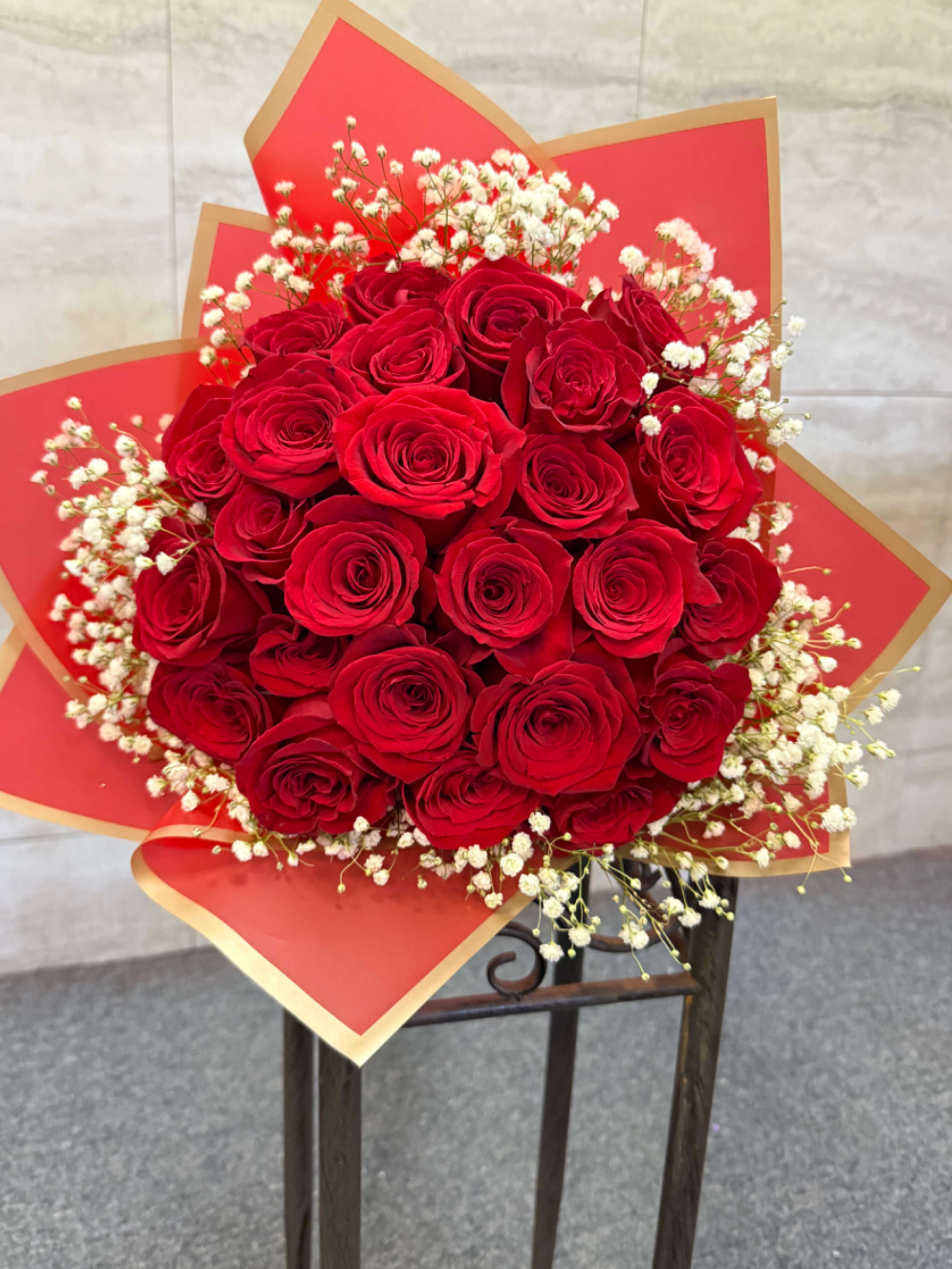 Round bouquet of red roses with white filler flowers wrapped in red and gold paper.