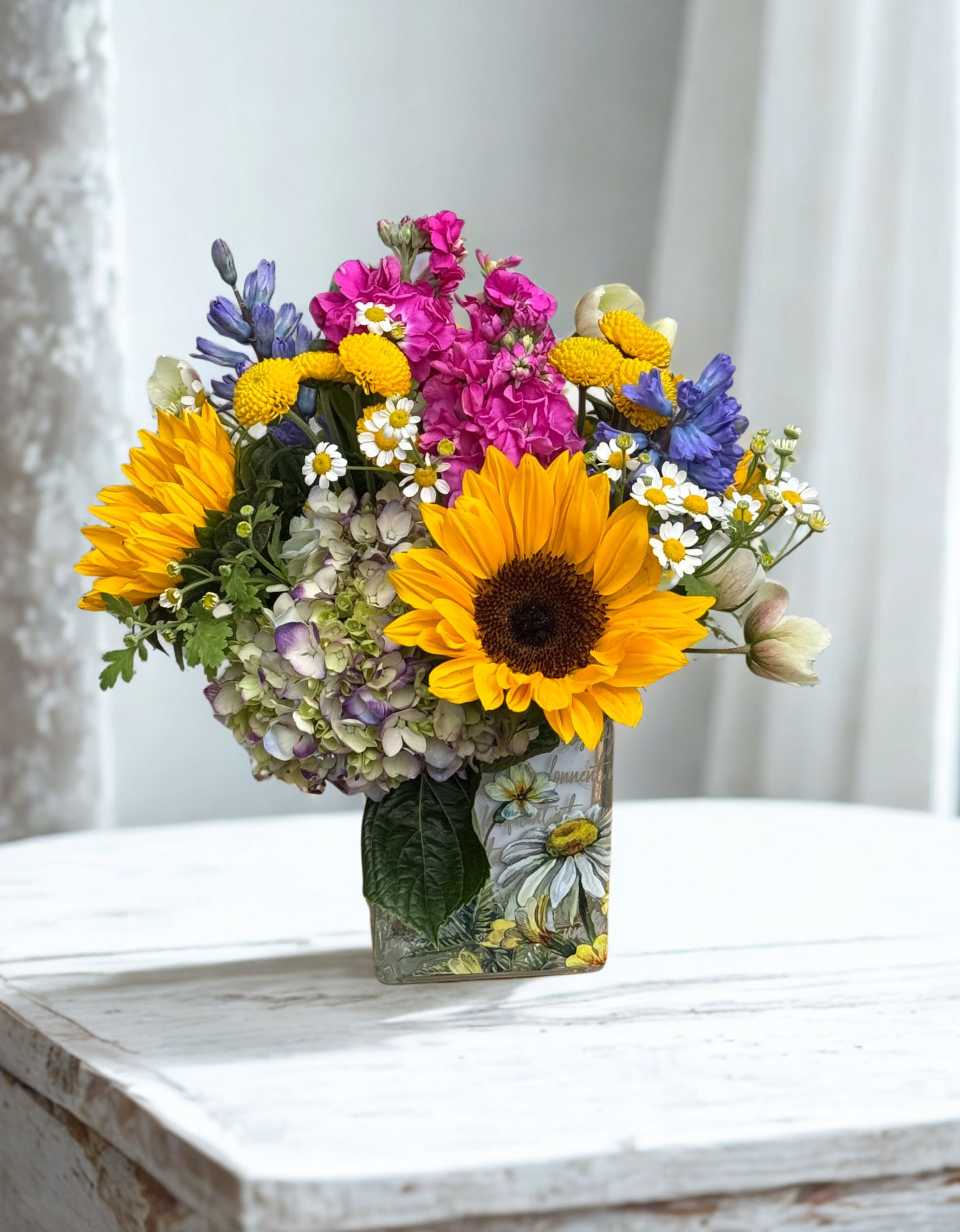 Busy Bee - This sunny arrangement features sunflowers, stock, hydrangea, buttons and hyacinth arranged beautifully in a cube vase.